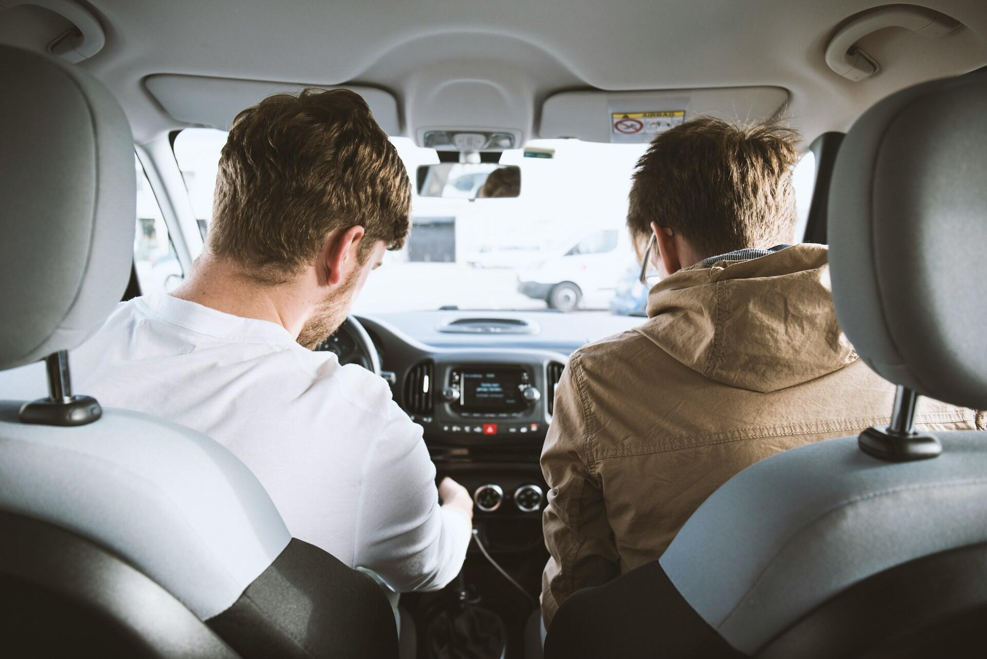 Two men sitting in the front seats of a car, viewed from the back seat, with one in a white shirt and the other in a brown jacket.