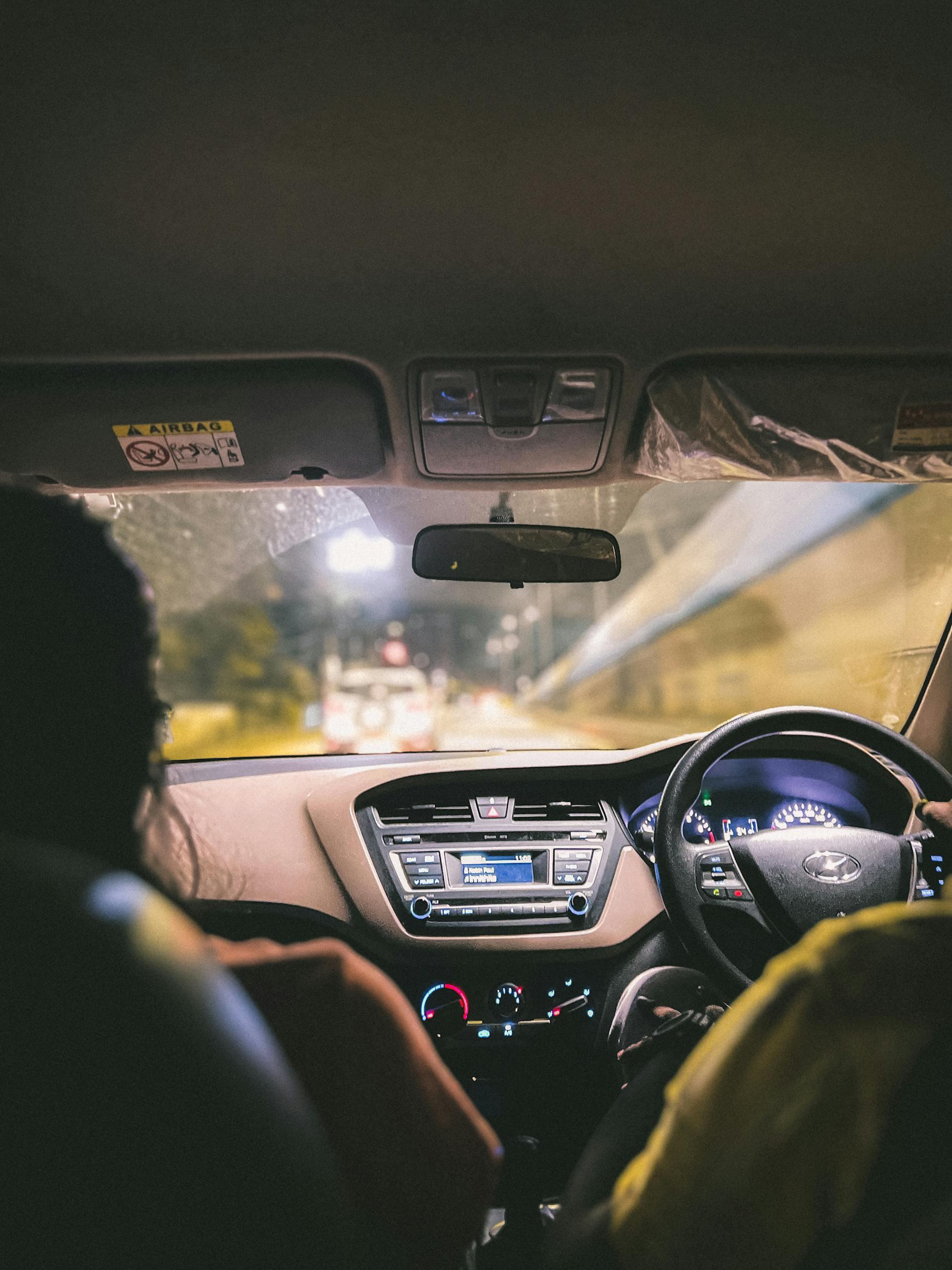 Interior view of a car at night with a driver and passenger, dashboard lights on, and a blurred road visible through the windshield.