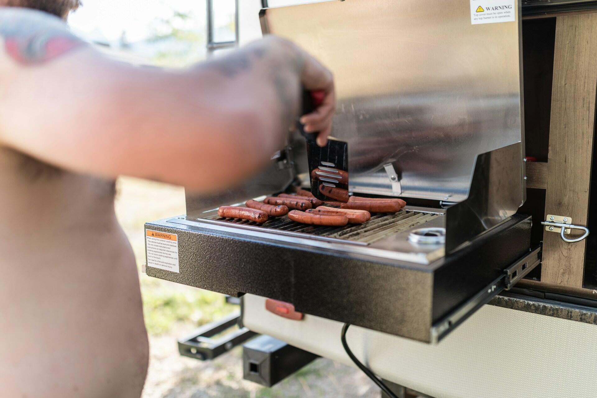 A person tends to sausages on a silver electric grill mounted on the back of a camper.
