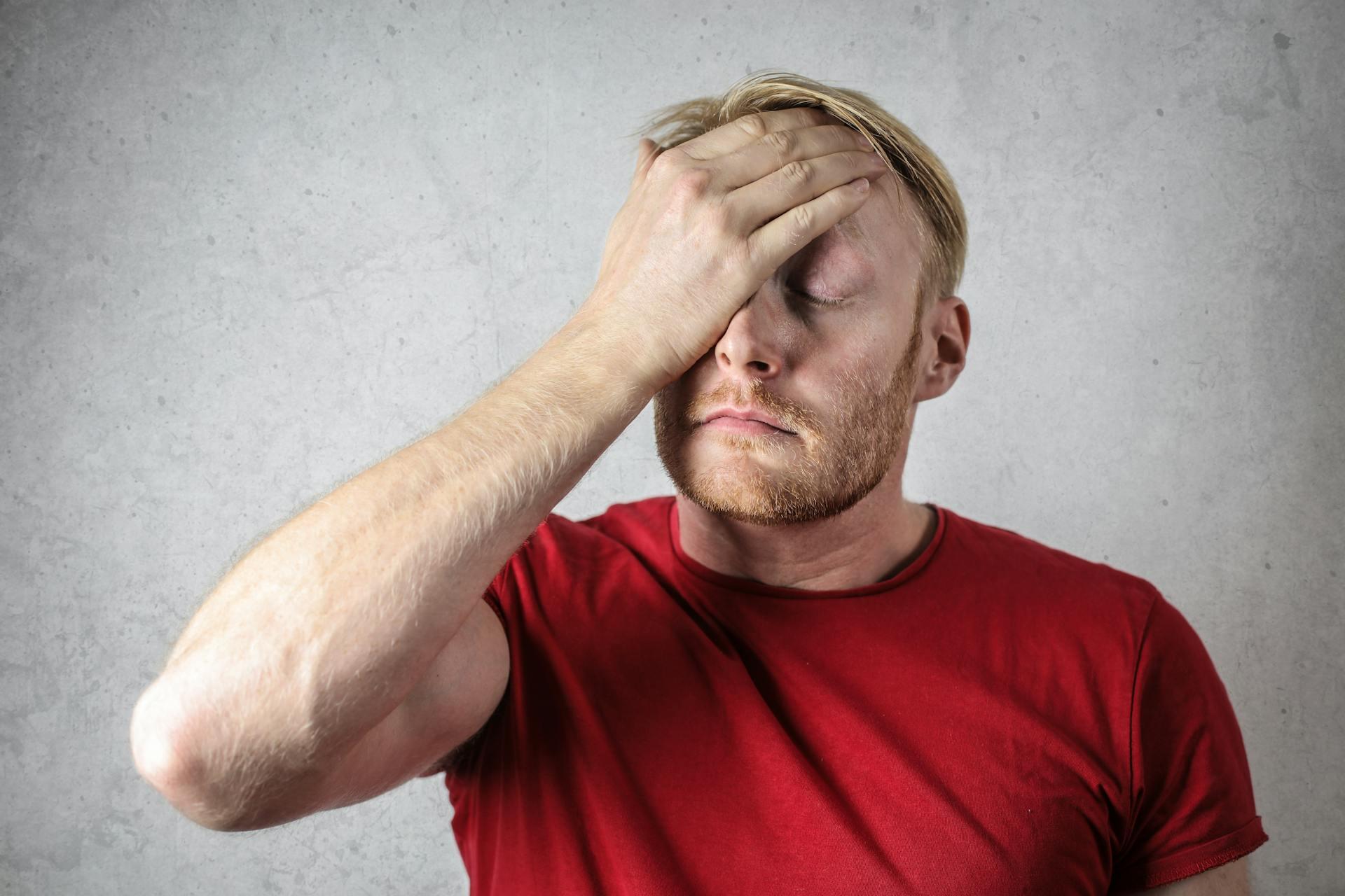Man in red shirt holding his head with one hand against a gray textured background.