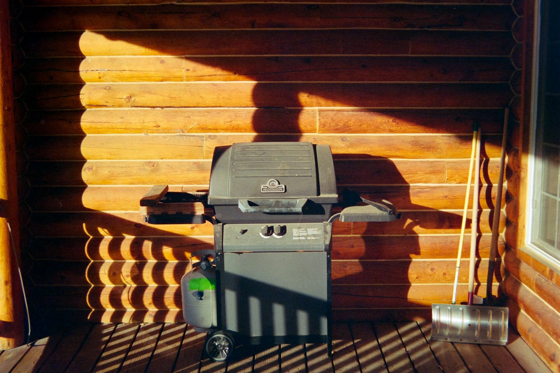 A grey two-burner gas grill with closed lid in front of a wooden wall on a porch on a sunny day.