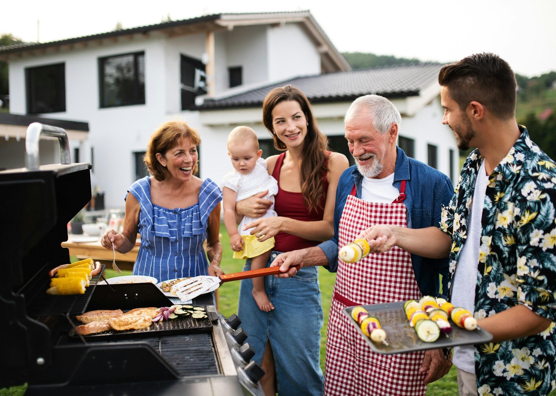 A group of people around a barbecue grill. A woman holds a baby while an older man works the grill.