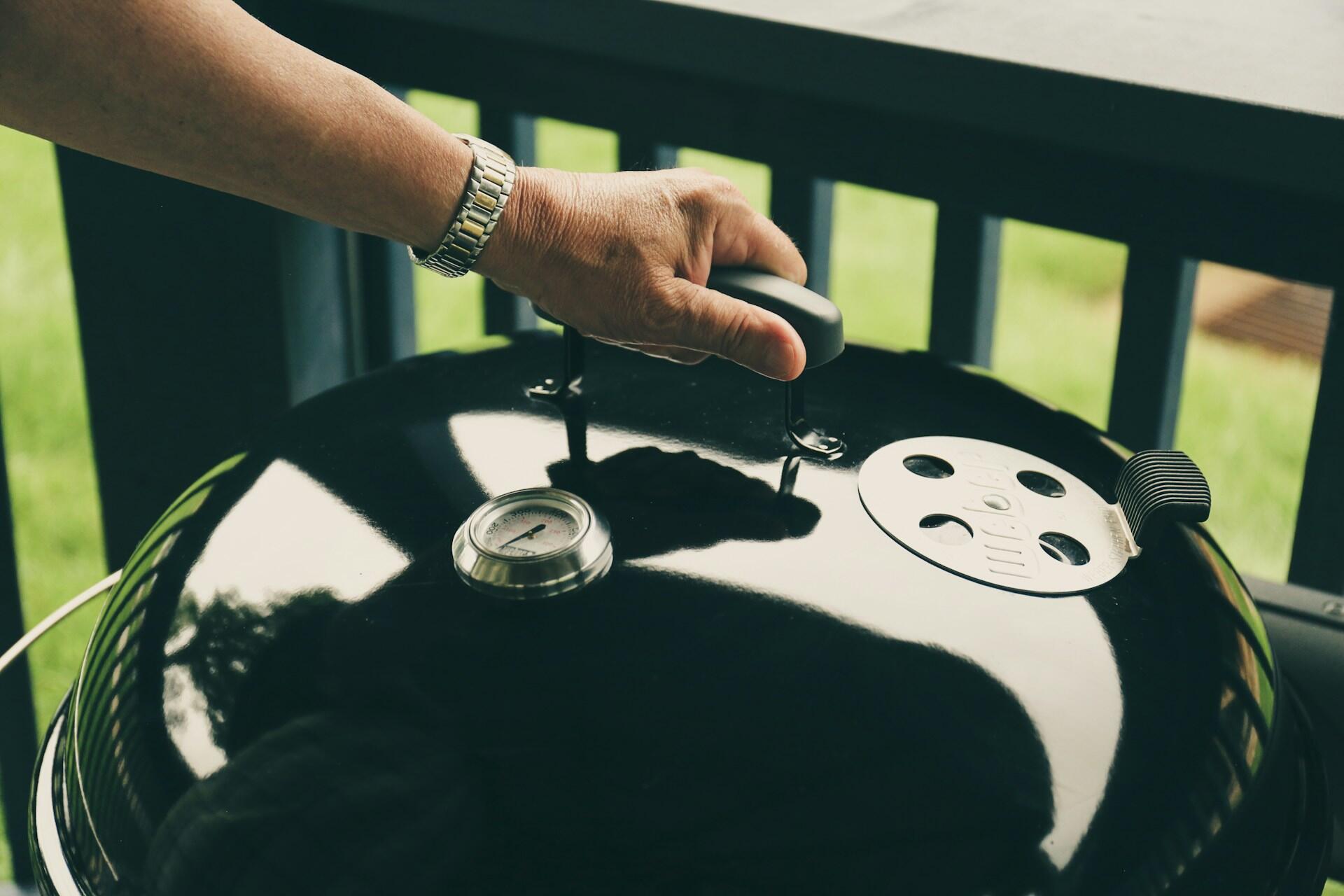 A person wearing a wristwatch with their hand on a black kettle grill's lid.