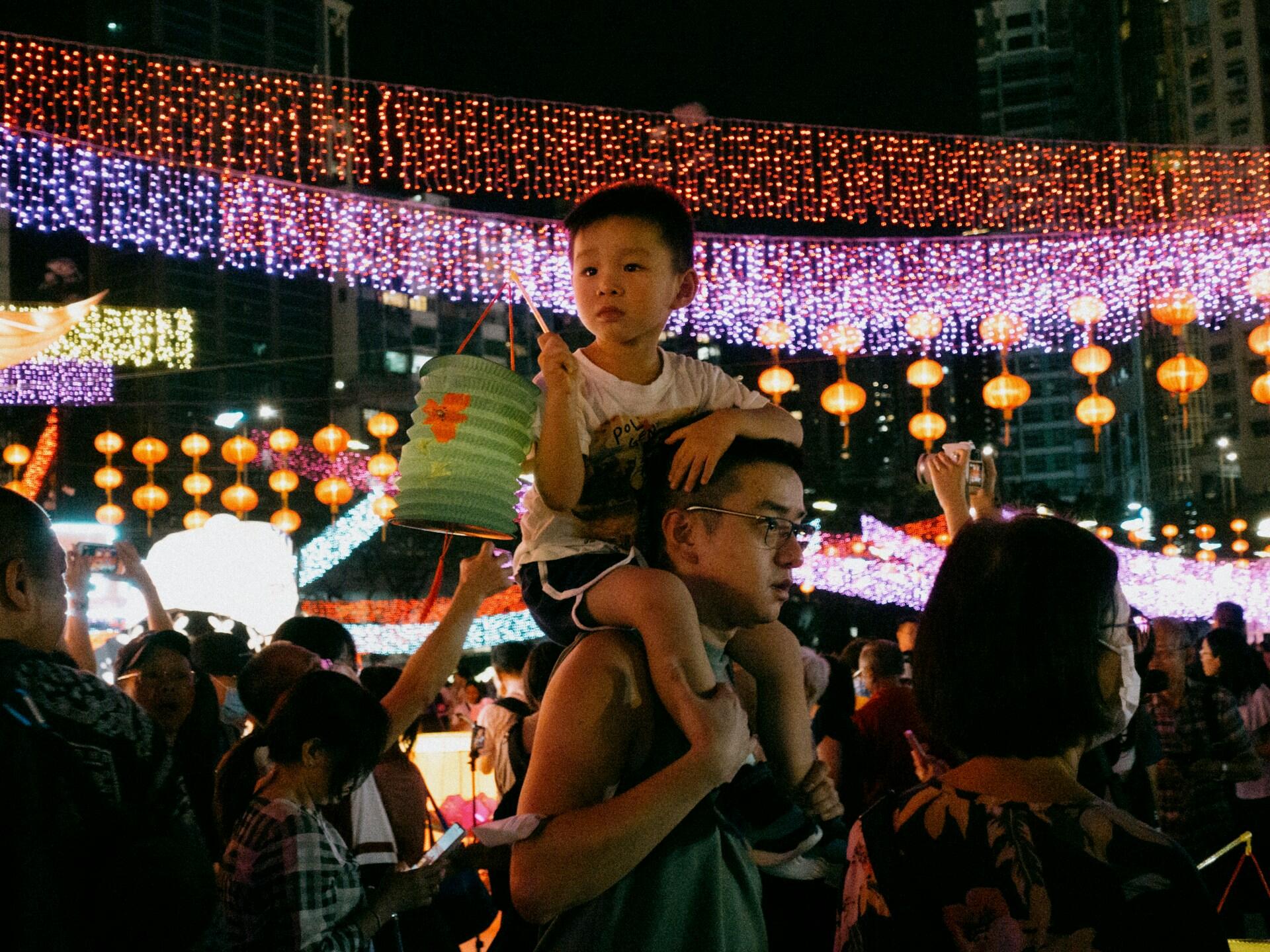 A child atop a man's shoulders holding a green paper lantern at night in a crowded place.