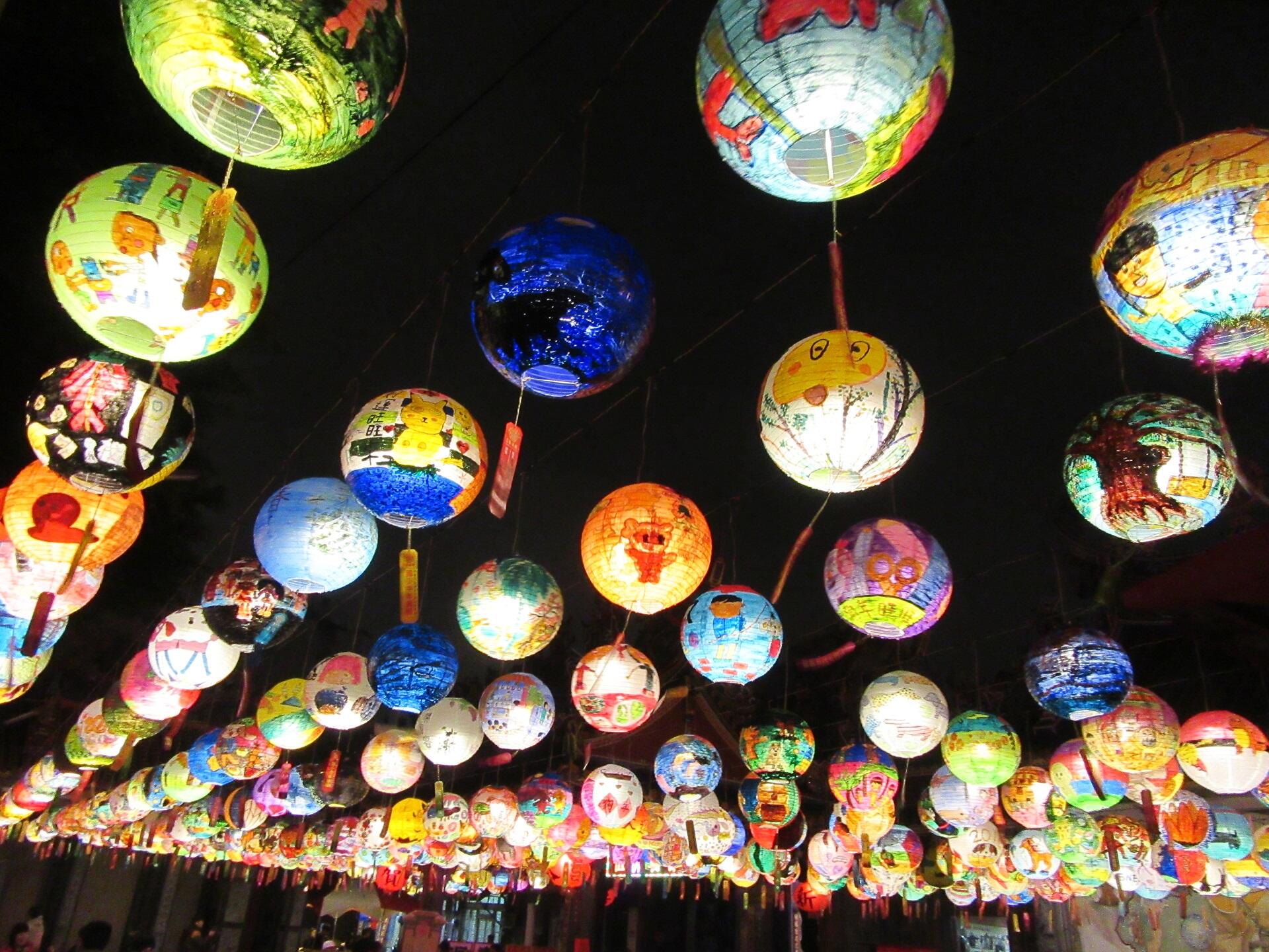 A display of colourful, painted lanterns with diverse, artistic designs hanging as a canopy against the night sky. 
