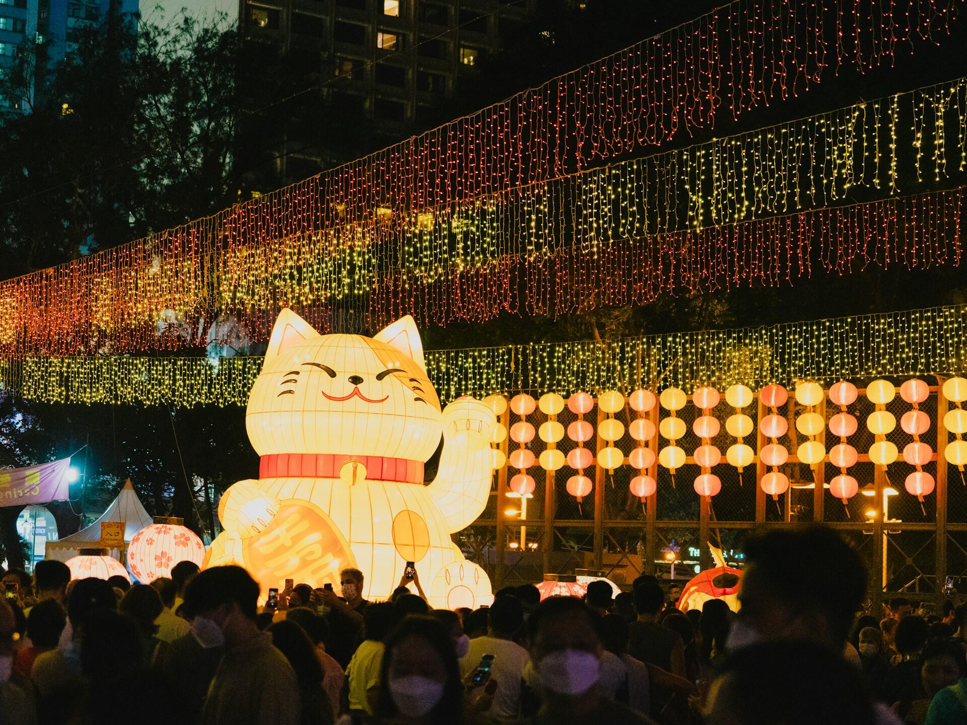 A lantern installation featuring a large cat lantern lit up at night, with many people walking through. 