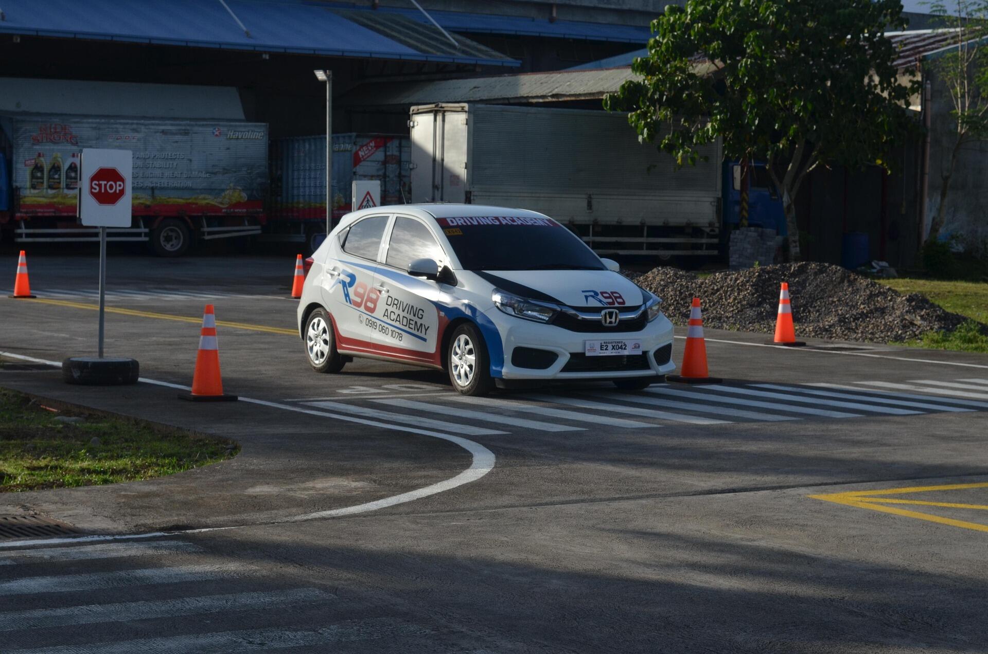 A white R98 Driving Academy hatchback is driving slowly over a marked crosswalk on an asphalt lot, surrounded by orange traffic cones and a stop sign.