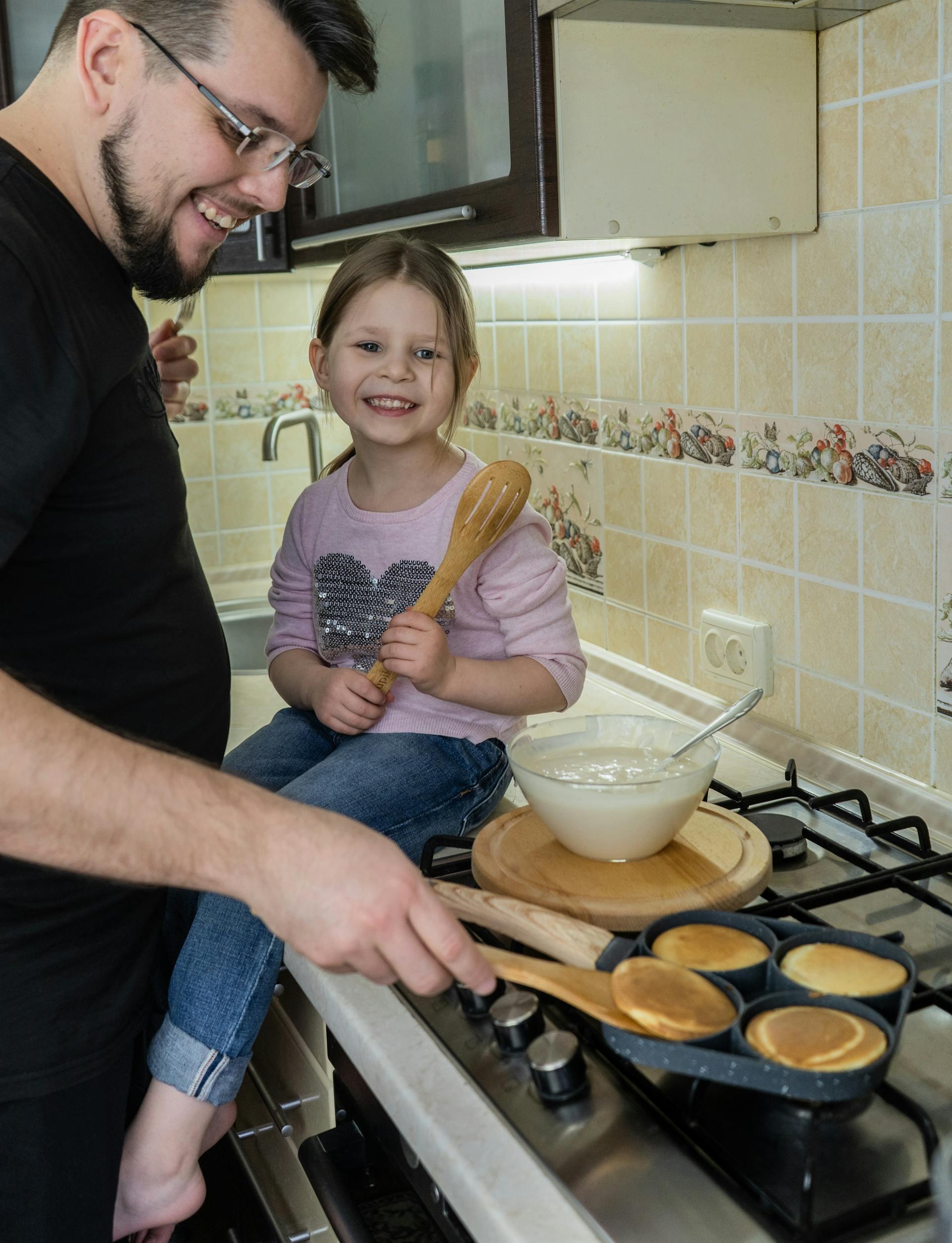 Smiling man and young girl cook pancakes together on a stovetop in a tiled kitchen.