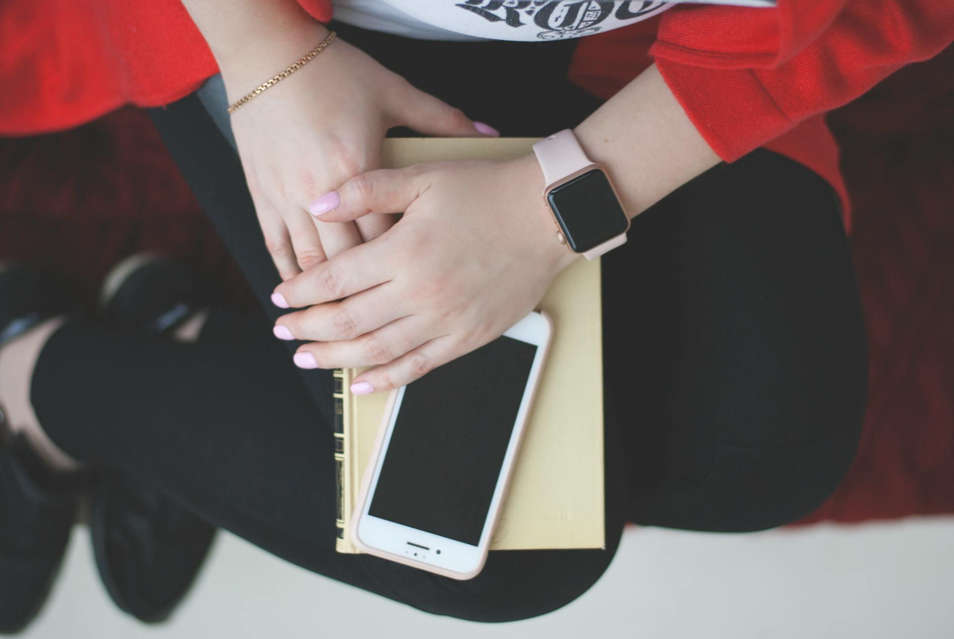 Woman wearing a red top and black pants sits while resting hands with pink nail polish and a smartwatch on a yellow book with a white smartphone placed on top.