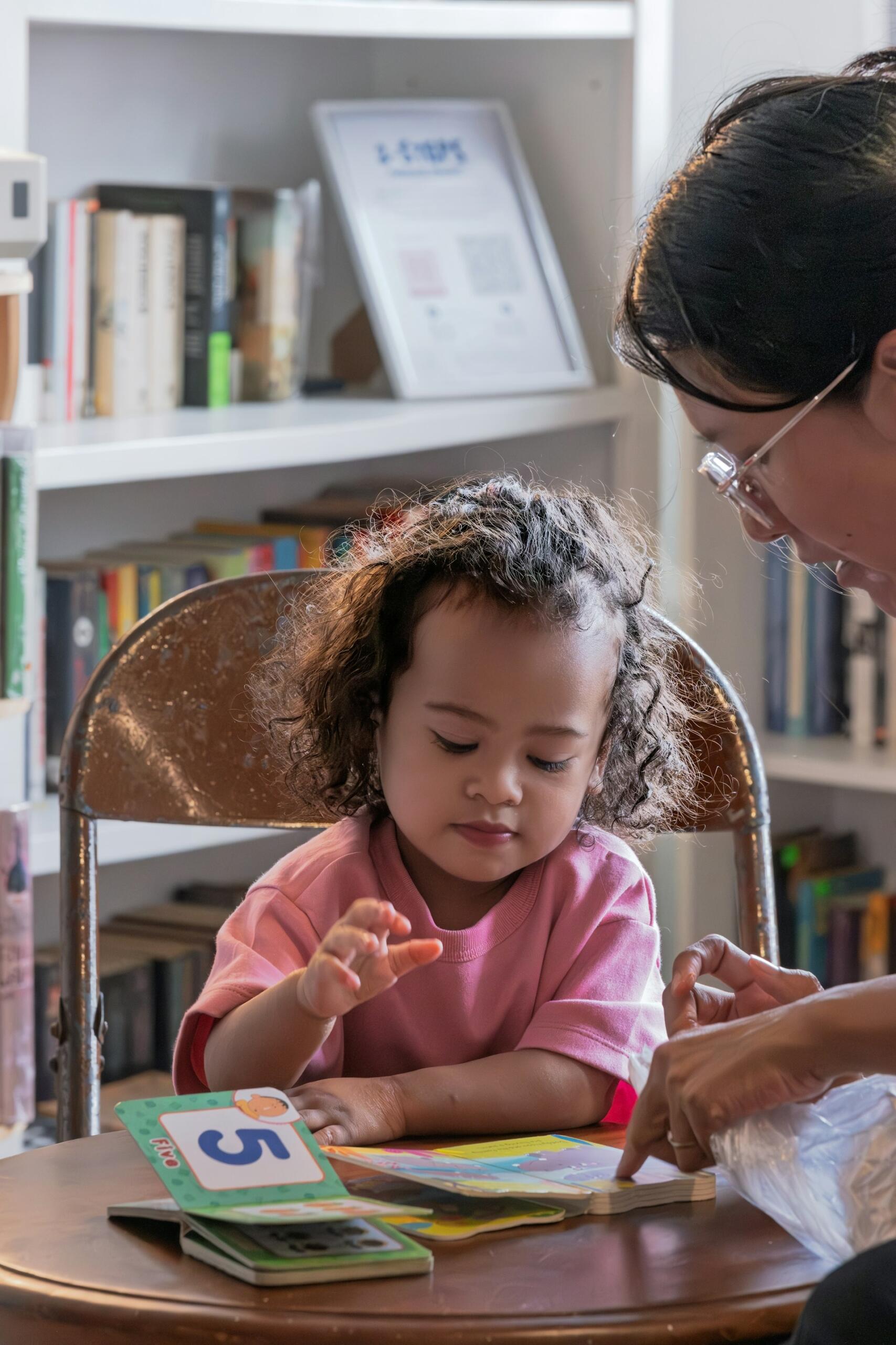 A young child in a pink shirt sits at a wooden table learning numbers with an adult in front of a blurred bookshelf.