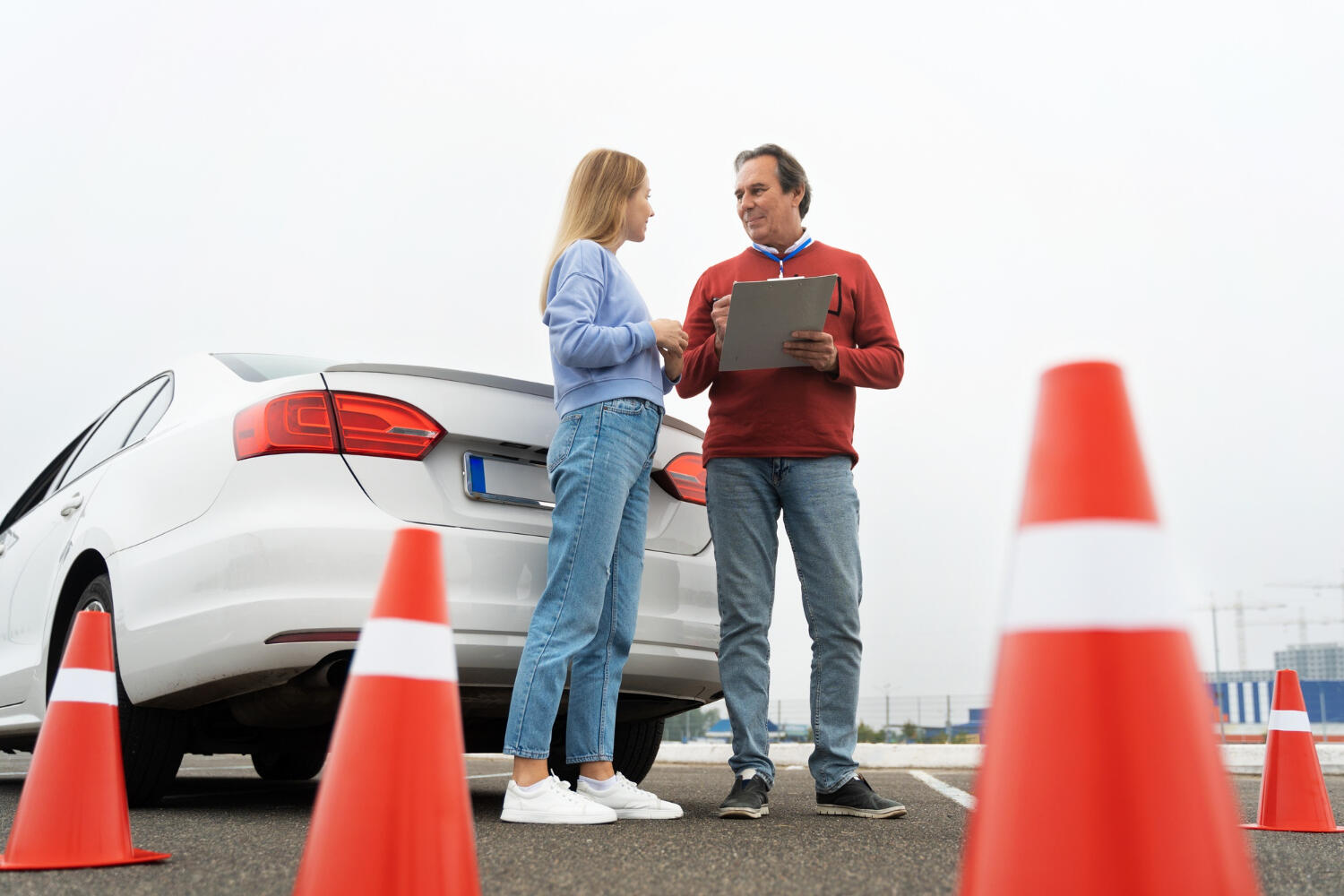 Two people standing near a white car in a parking lot, surrounded by orange traffic cones, with one holding a clipboard.