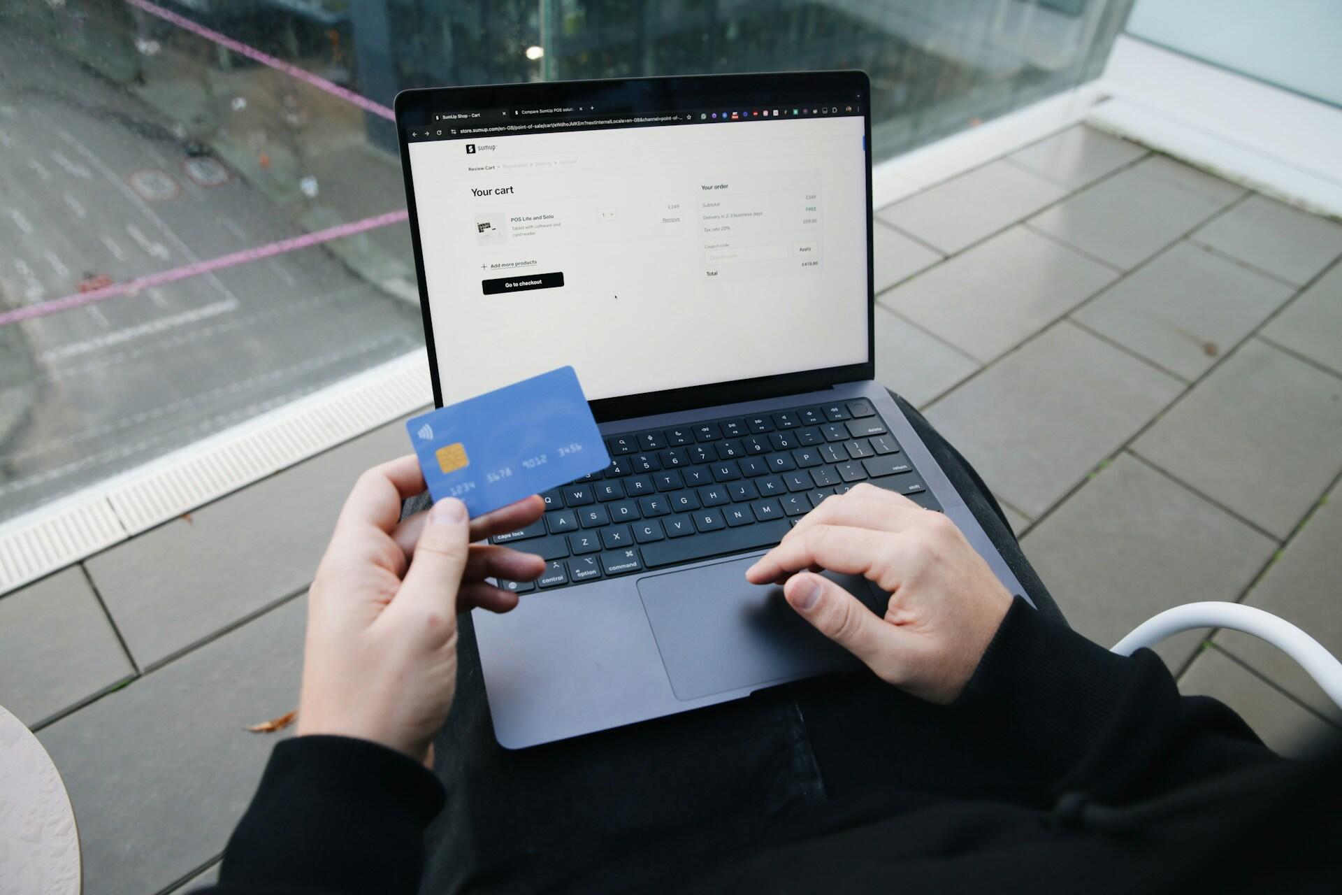 Person holding a blue credit card and using a laptop to complete an online purchase on a website's shopping cart page.