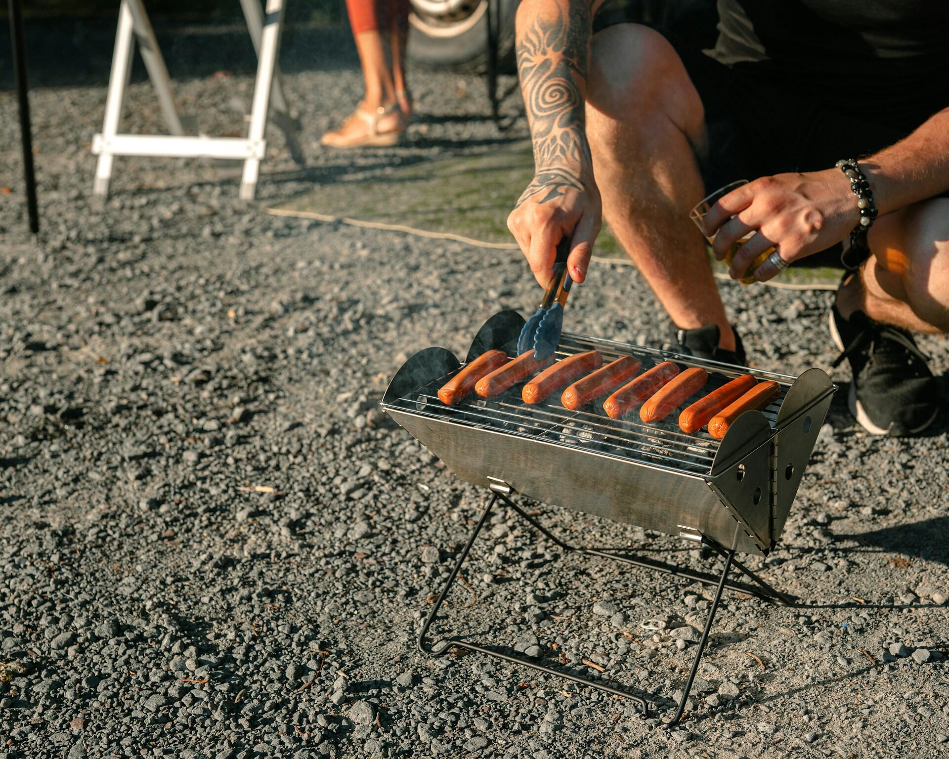 A small, silver, v-shaped portable grill with a man tending to the sausages grilling on it.