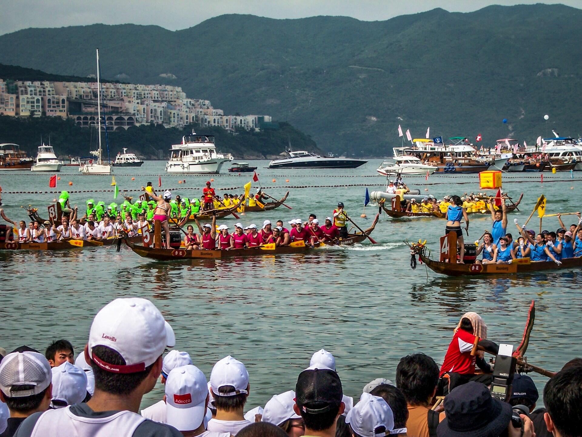 Dragon boat race with teams in jerseys paddling on calm water, surrounded by spectators in white caps and yachts against a hilly backdrop.