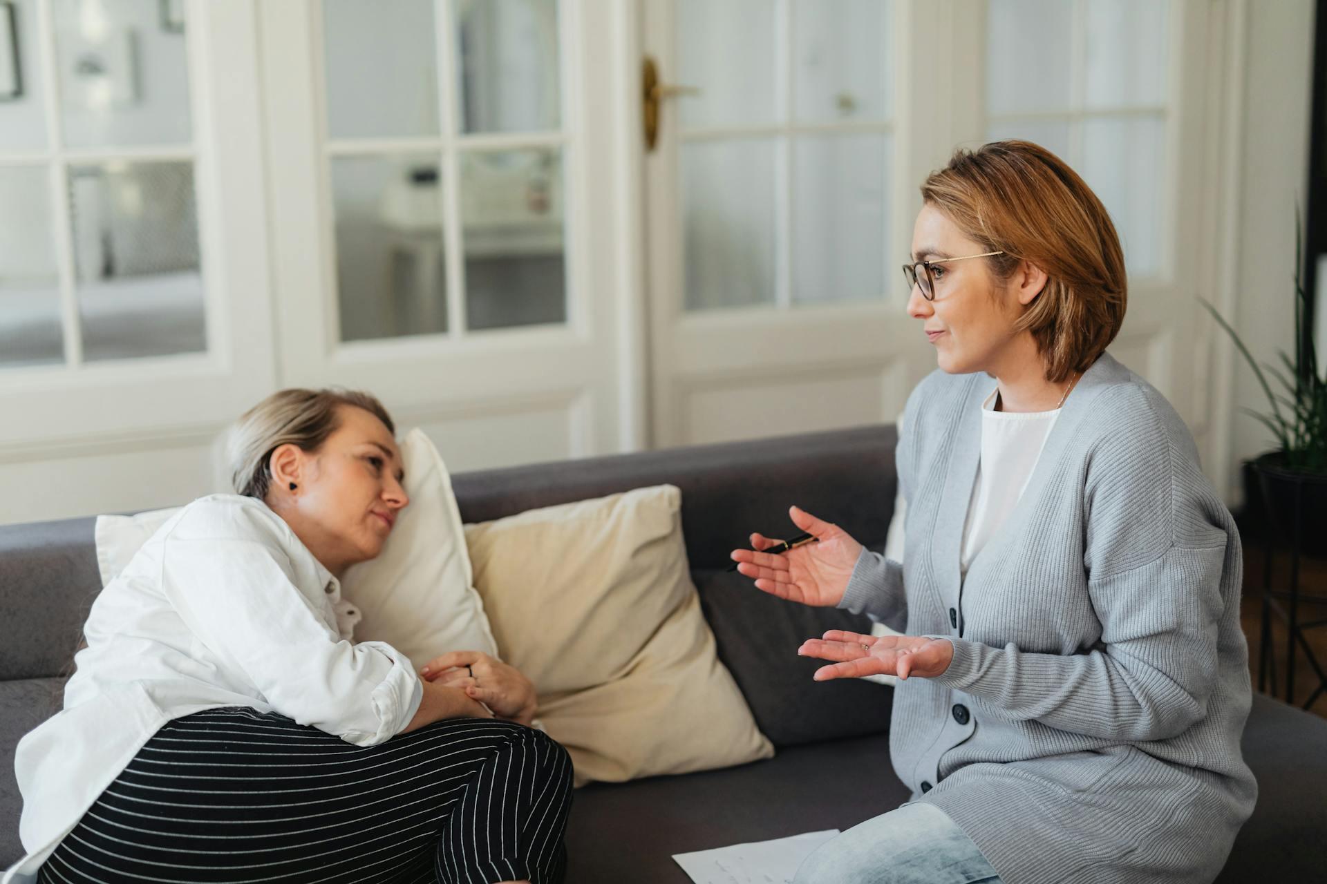 Two women having a serious conversation on a couch in a bright, cozy room with glass-paneled doors in the background.