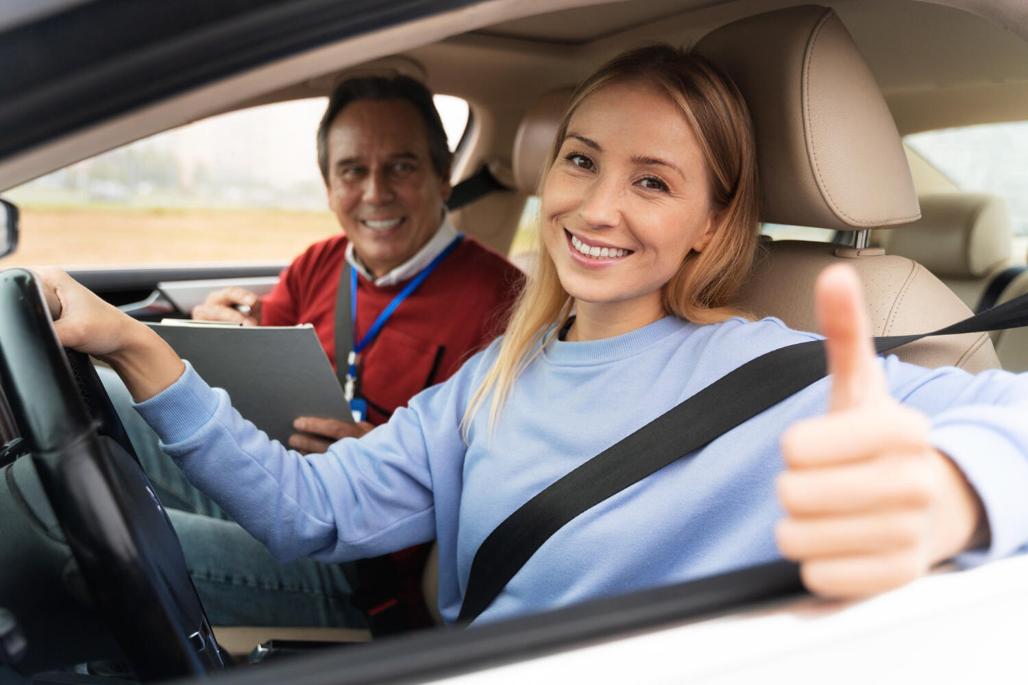 Young woman in blue sweater giving thumbs up while sitting in car with driving instructor holding clipboard.