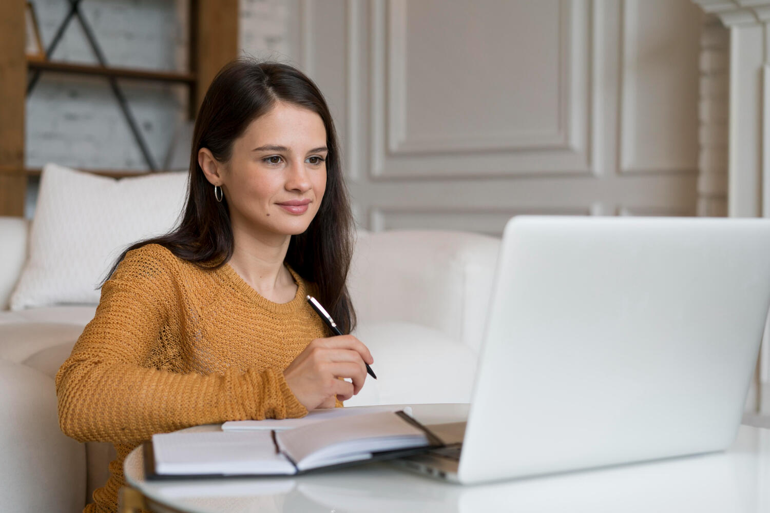 Person in a mustard sweater sitting at a table with a laptop and notebook, writing with a pen in a cozy indoor setting.