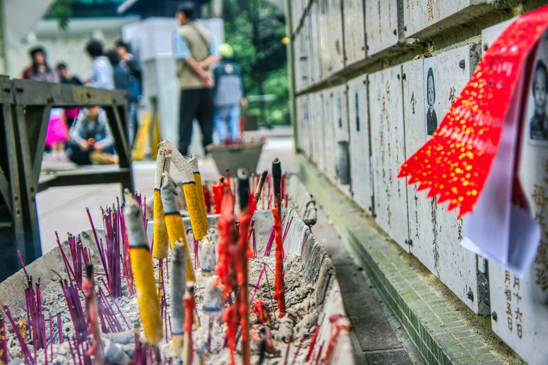 Incense sticks in various colors burn in a metal tray, set beside a textured columbarium wall with red paper decorations, and a group of people in the background.