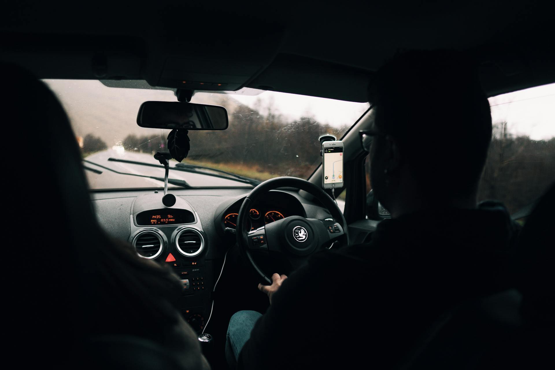View from the backseat of a car showing the driver navigating with a mounted phone displaying a map on a wet, overcast day.