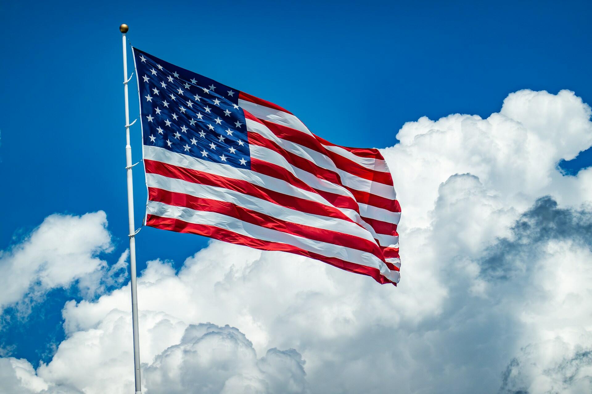 American flag waving on a flagpole against a bright blue sky with large white clouds.