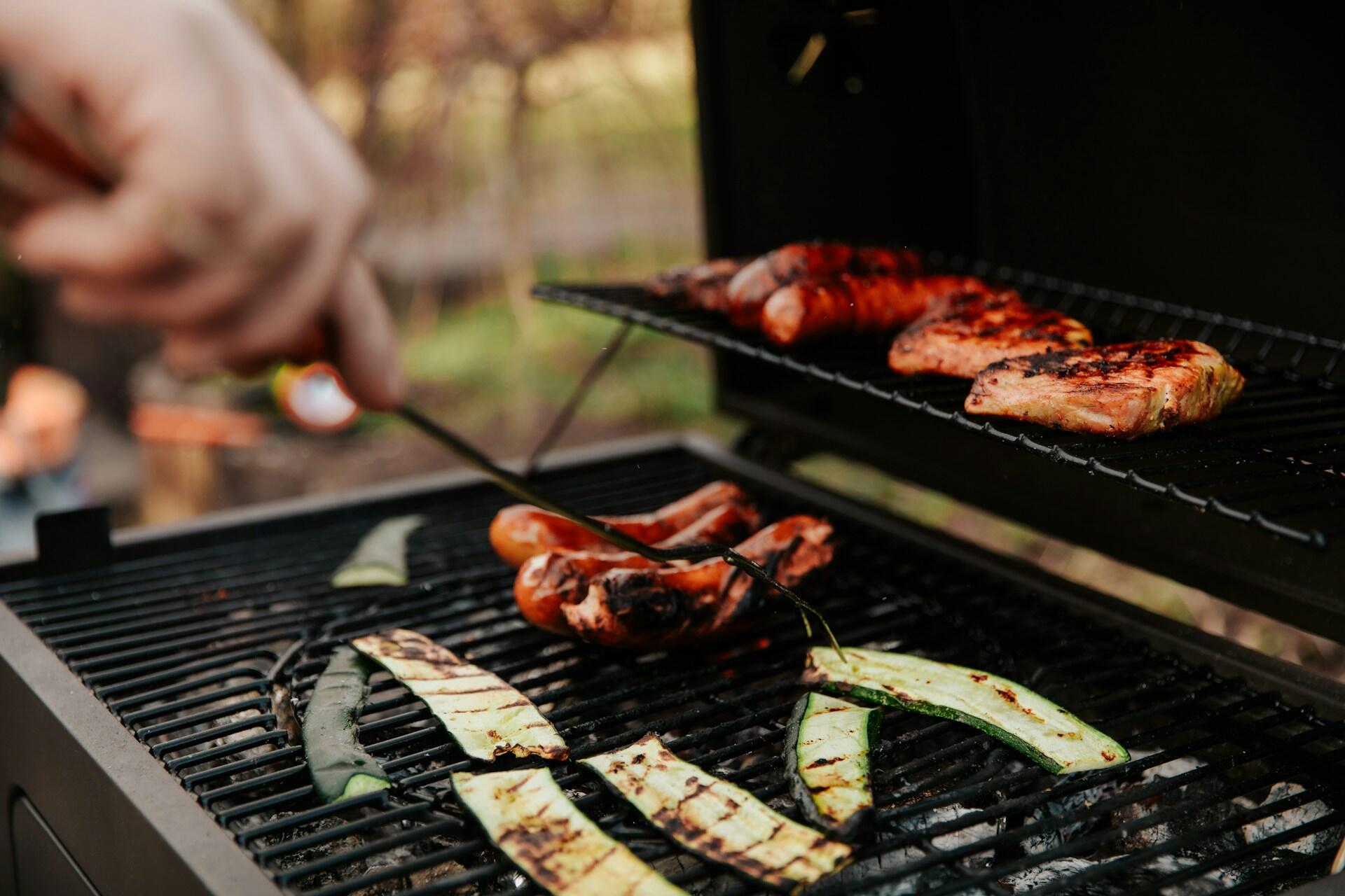 Hand grilling sausages, chicken, and zucchini slices on an outdoor barbecue grill with a fork.