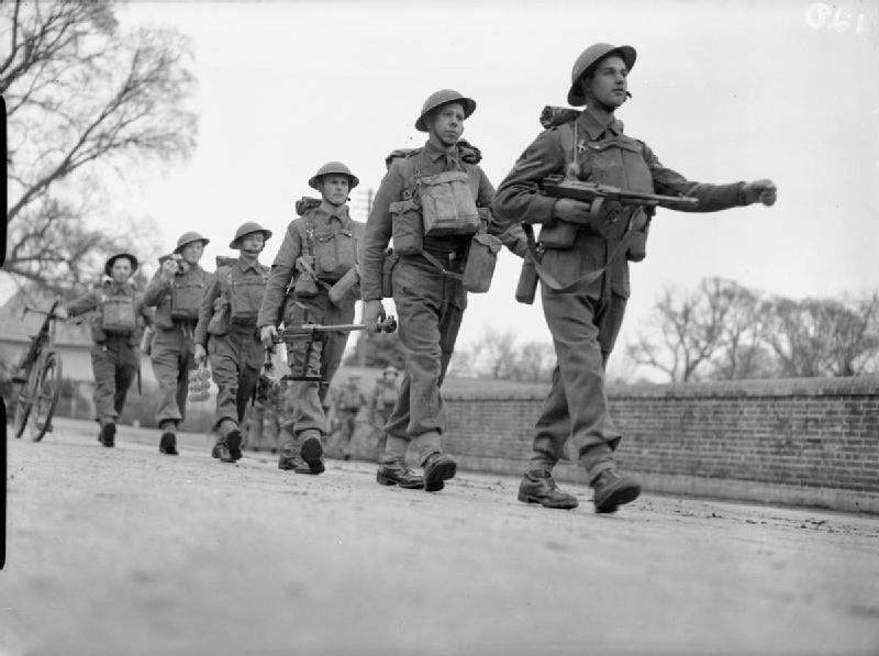 Black and white photo of soldiers in uniform and helmets marching in a line along a street with trees and a brick wall in the background.