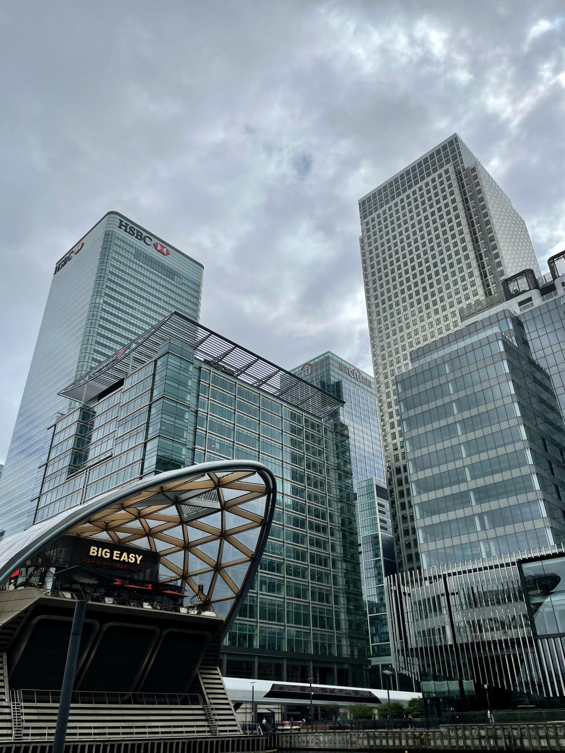 Modern glass skyscrapers, including the HSBC building, rise against a cloudy sky, with a distinctive curved structure in the foreground displaying