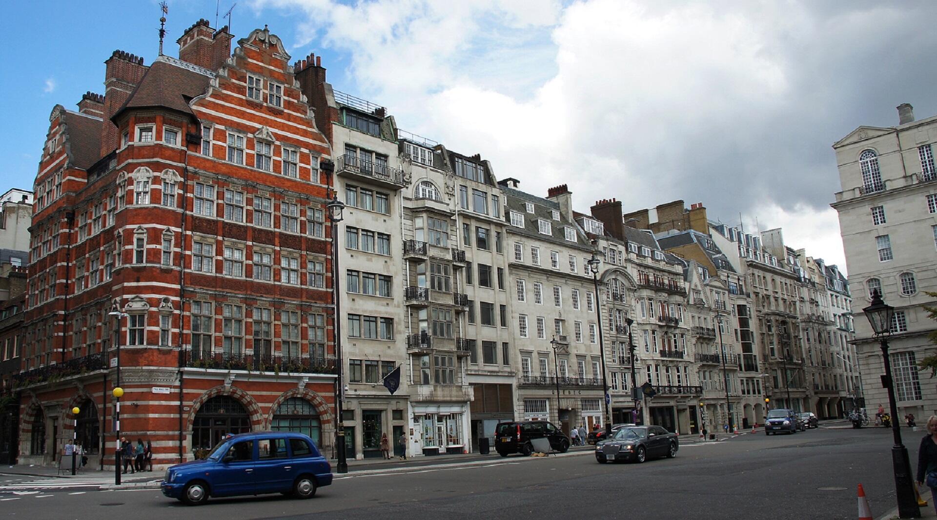 A row of tall buildings, one with a brick facade with others white-fronted, on a broad street with cars on it.