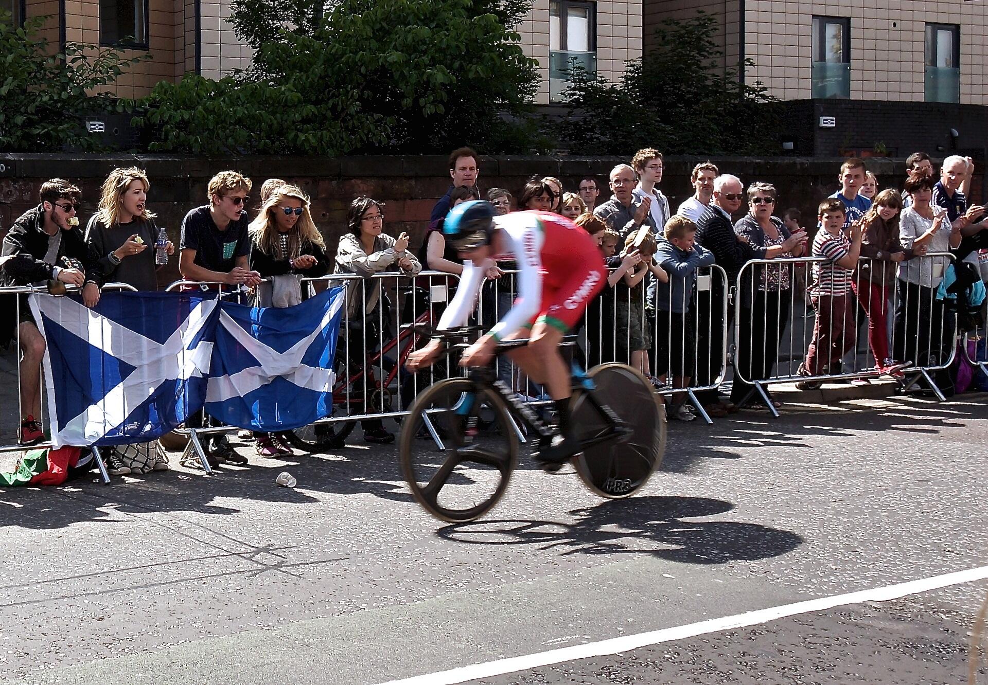 A cyclist wearing red and white rides in front of spectators behind barriers adorned with Scottish flags. 
