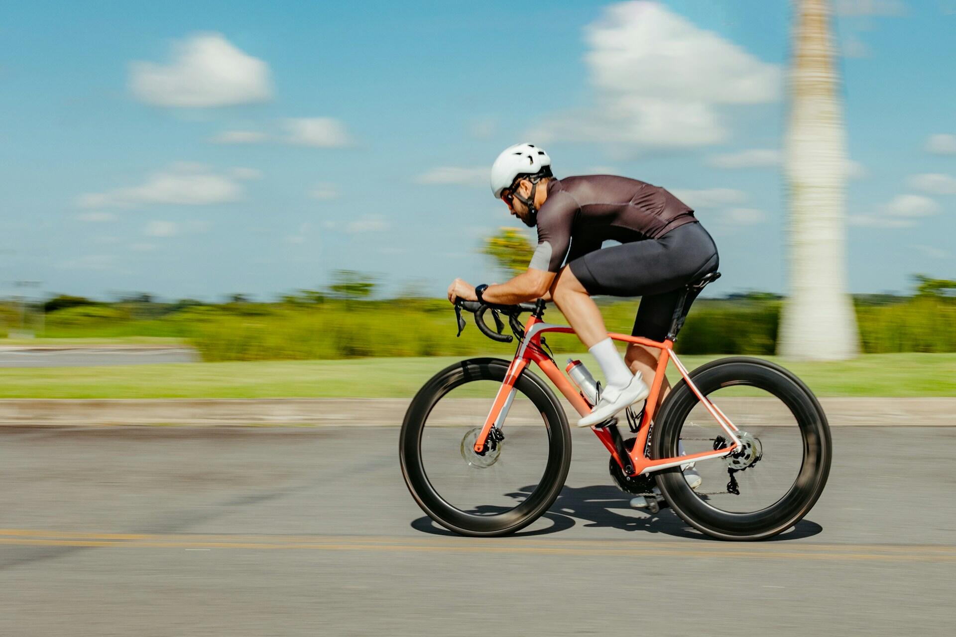 A cyclist wears a white helmet, black shorts and a black top while riding an orange bike on a sunny day.