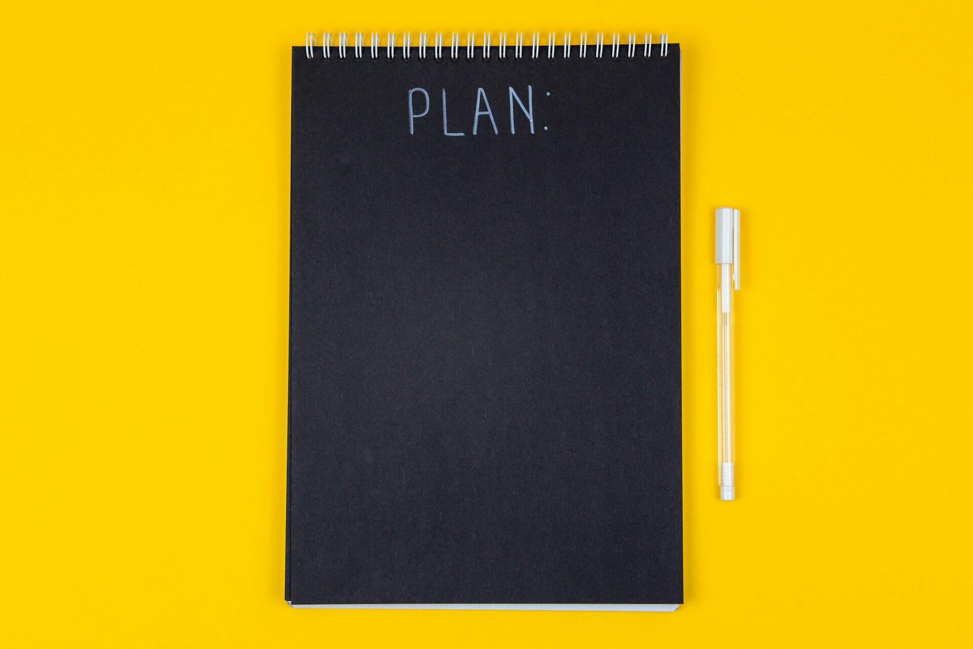 A black notebook with the word “plan” written on it and a pen on a yellow background
