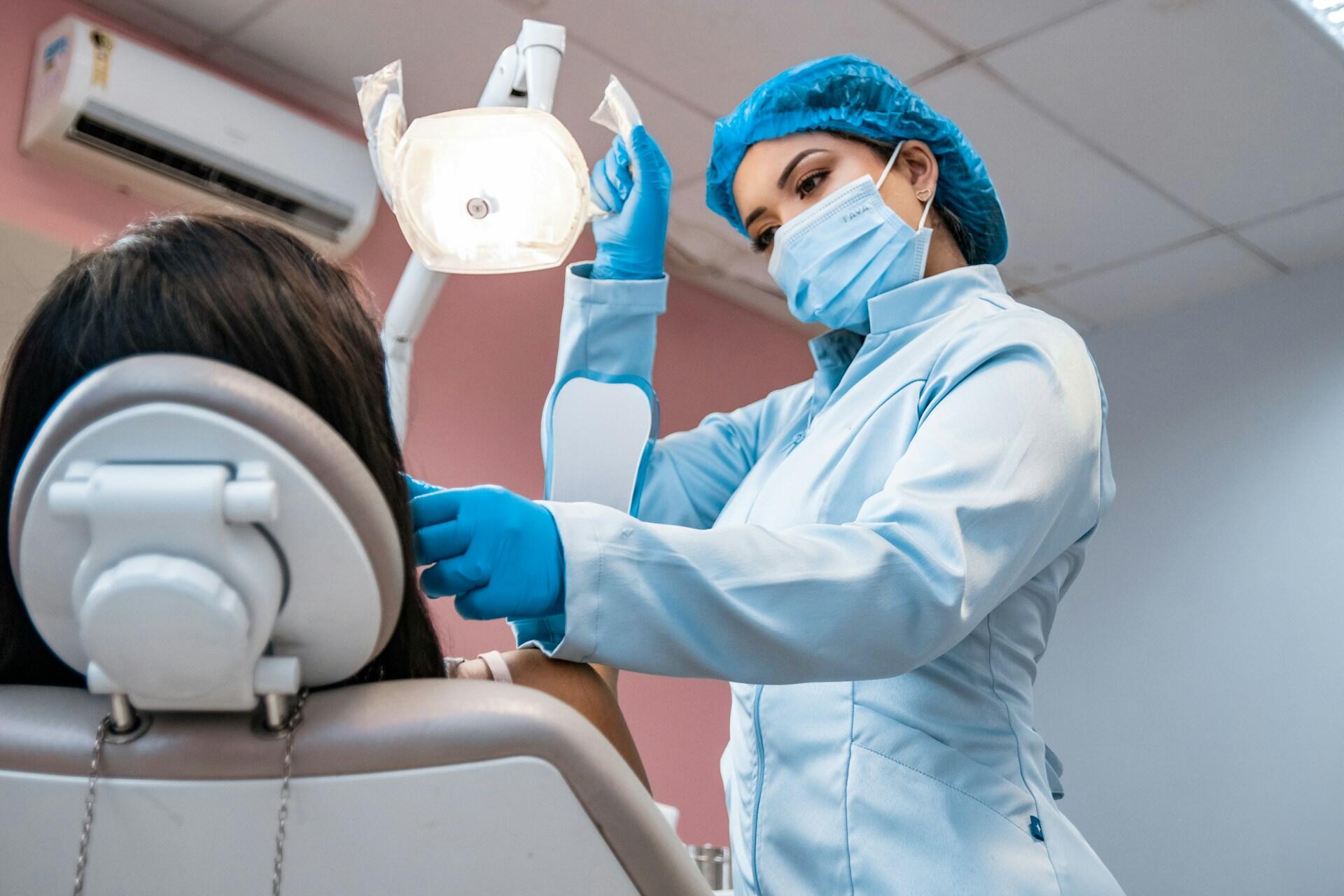 Dentist wearing a mask, gloves and blue cap treating a patient in a dental chair