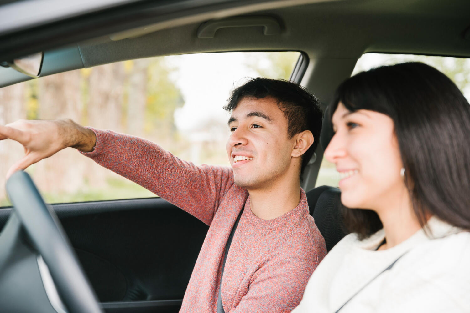 Two people sitting in a car, the driver pointing out the window while the passenger watches attentively.