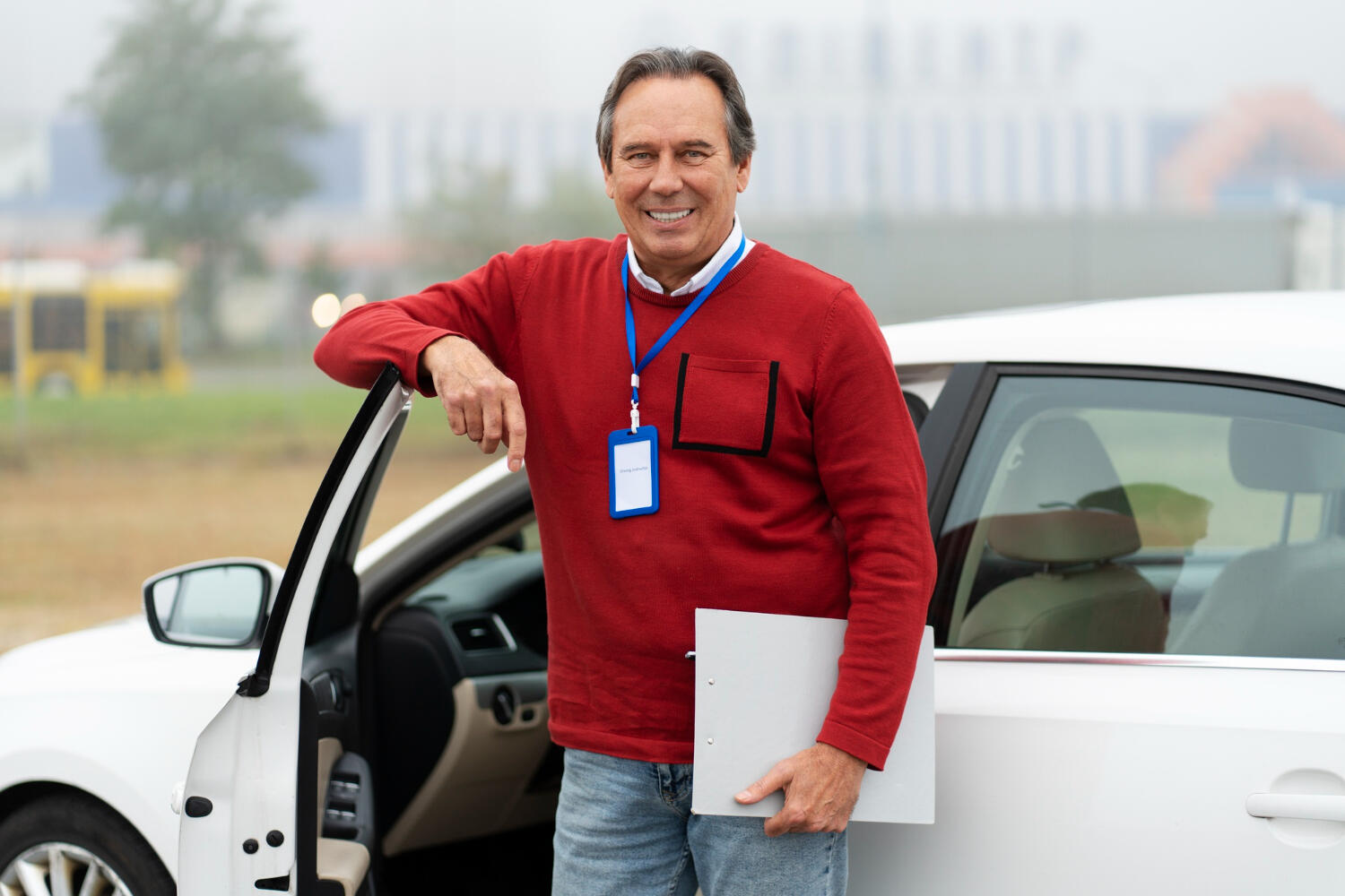 Man in red sweater holding clipboard, standing next to an open white car door in an outdoor setting.