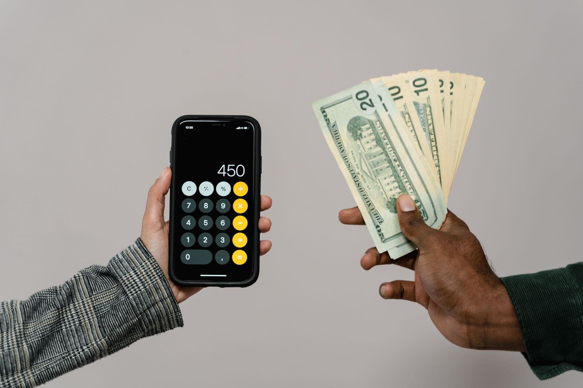 two hands, one holding a selection of dollars, the other a smartphone displaying a calculator