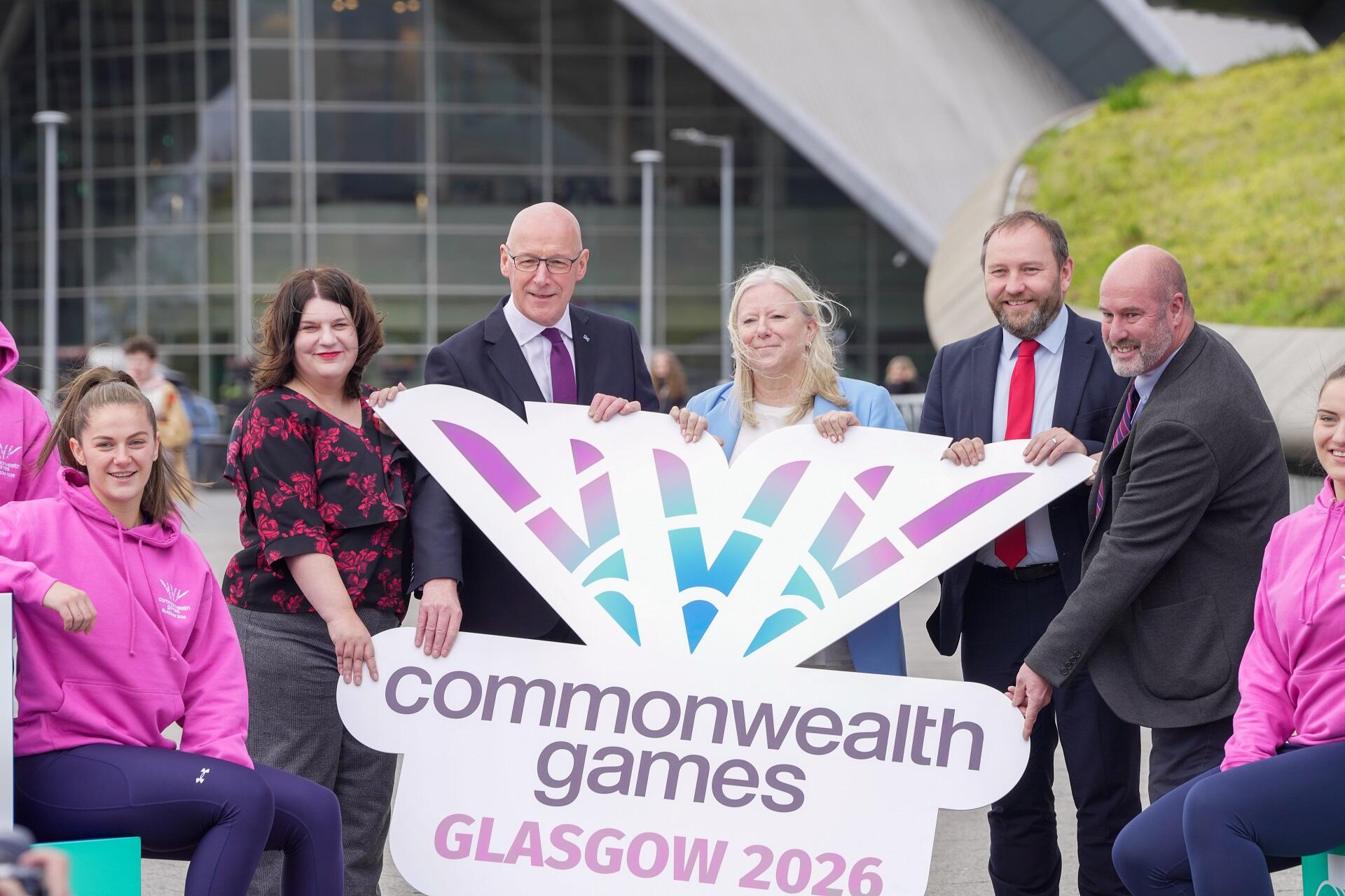 A group of people holding a colourful sign pose in front of a glass and steel building.