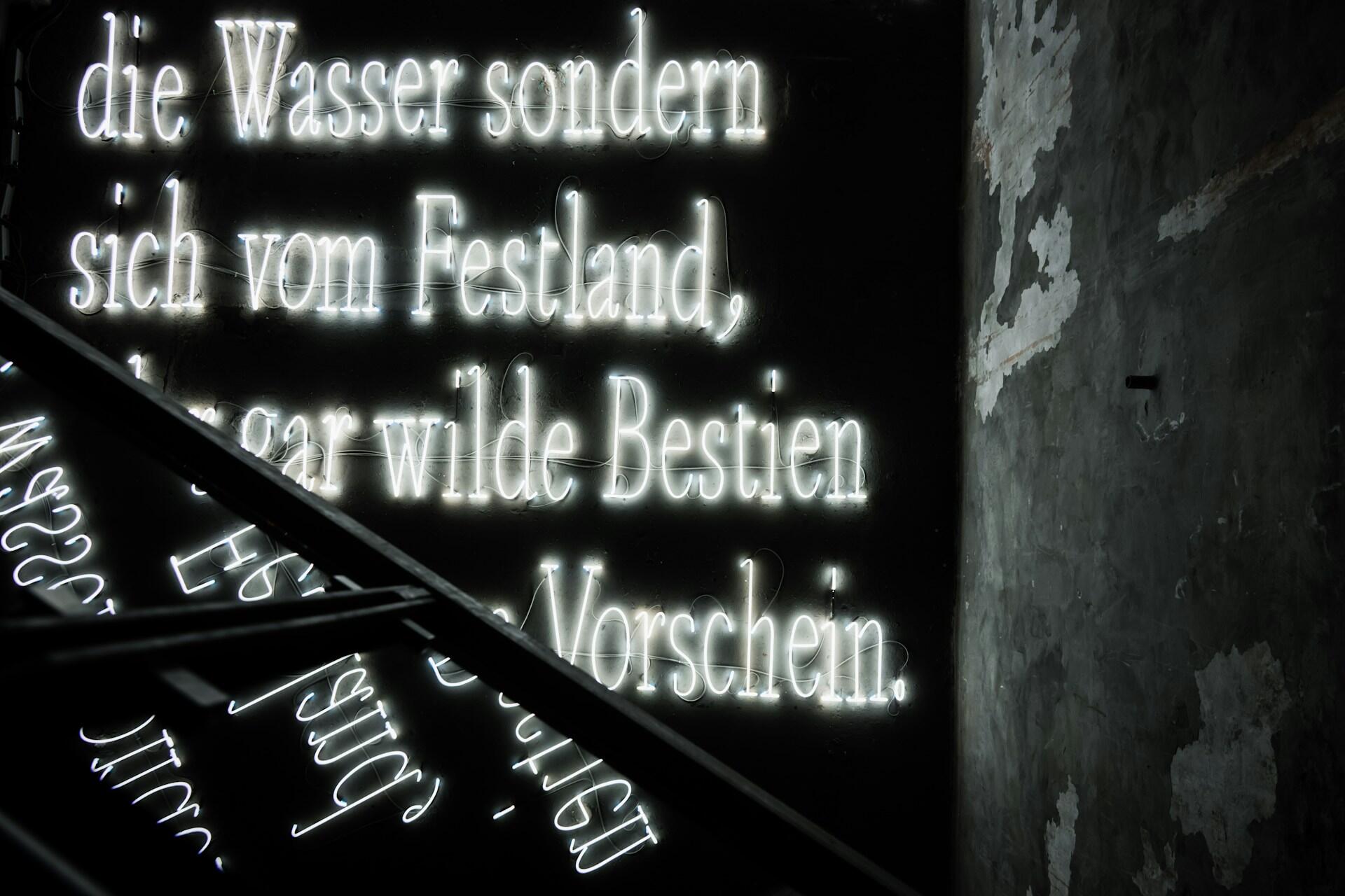 White neon German text on a dark wall beside a rough concrete surface.