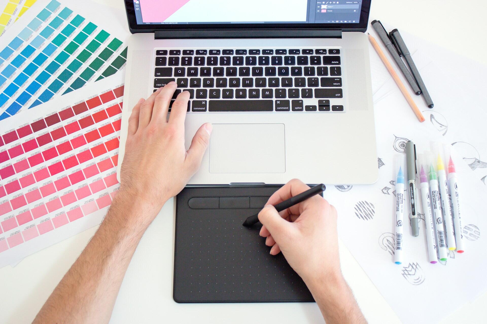 Graphic designer using a drawing tablet beside colour swatches, a laptop, markers, and sketches
