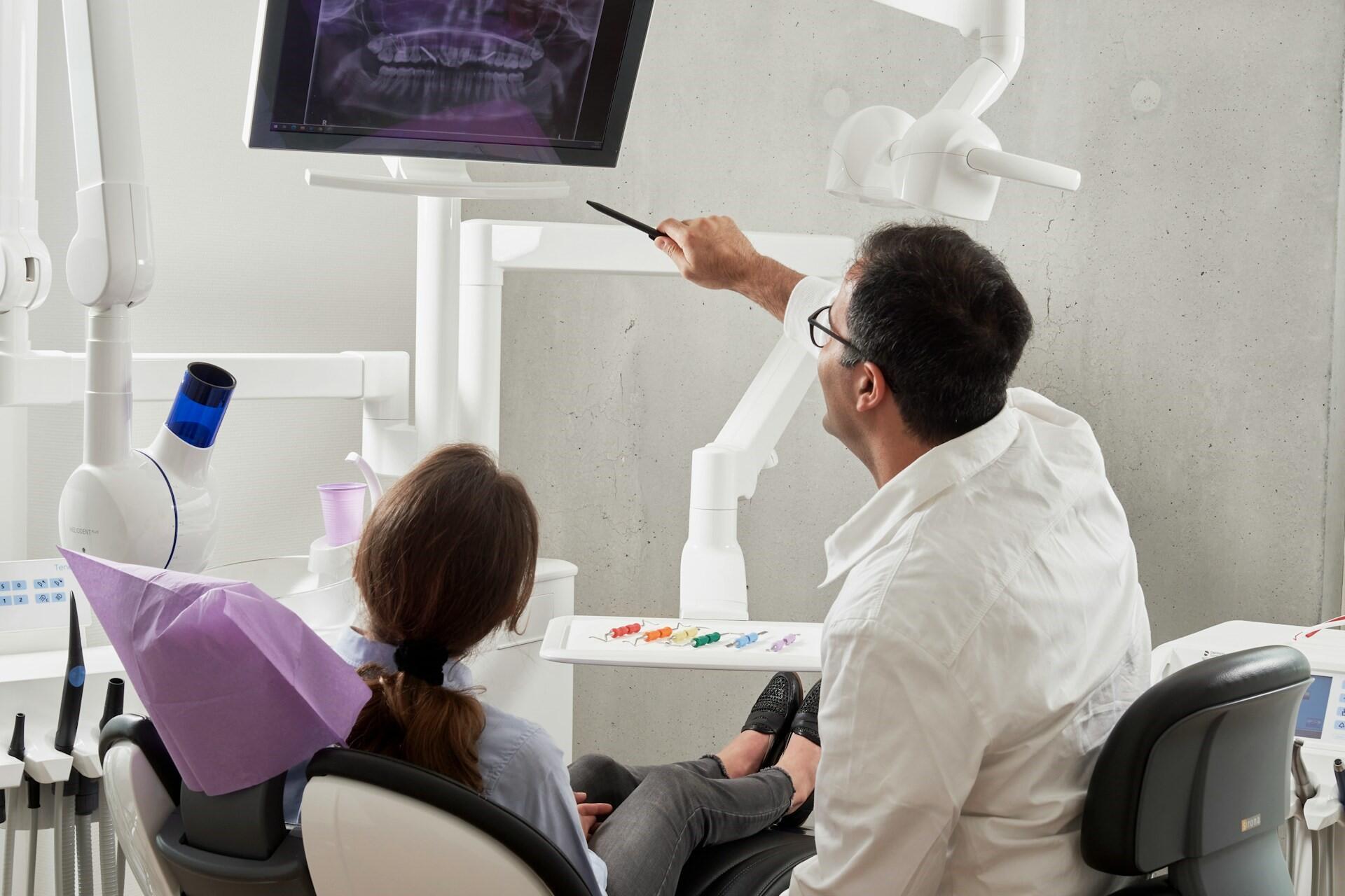 Dentist pointing at a dental X-ray on a screen while a patient sits in a dental chair