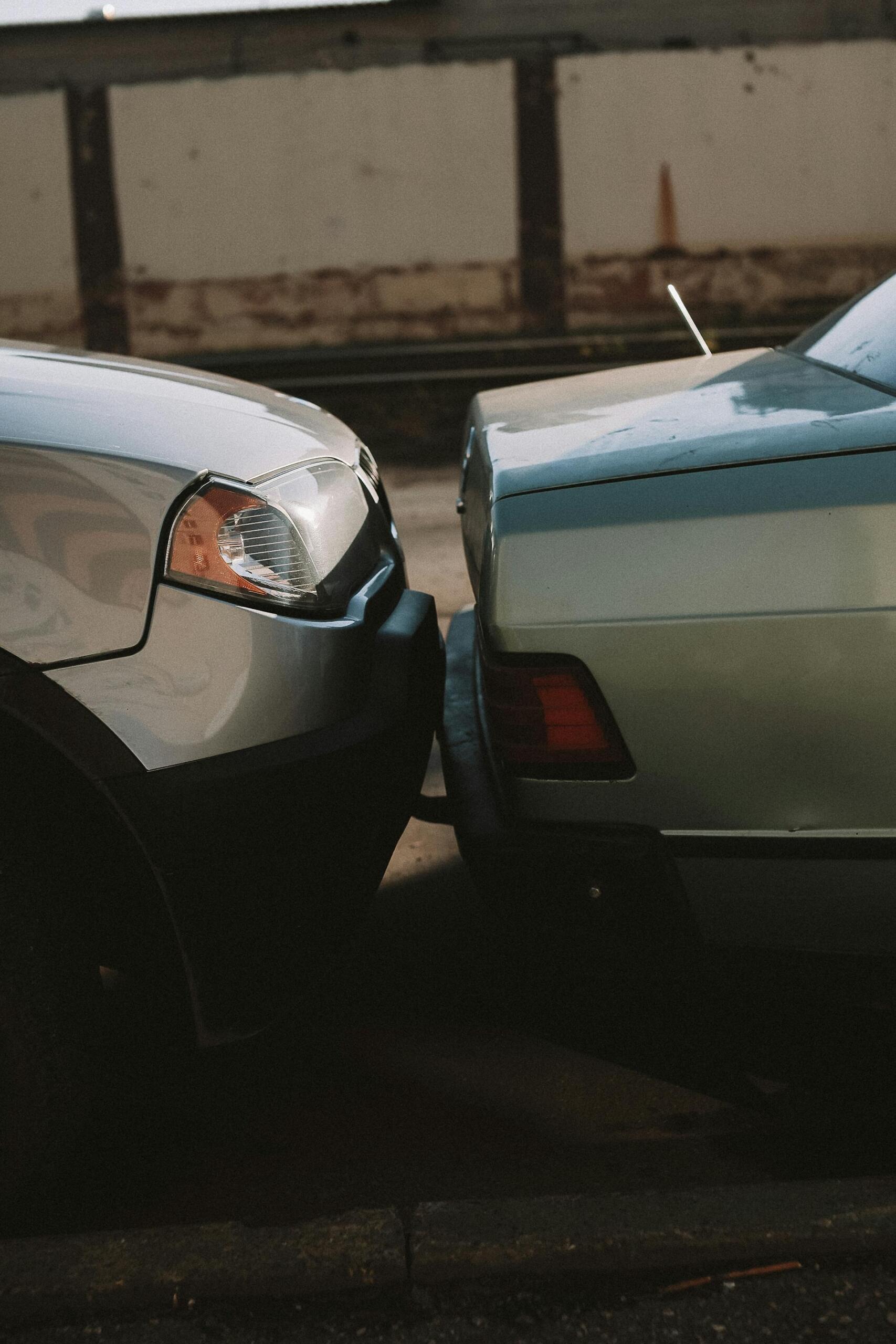 Two cars parked bumper-to-bumper in a dimly lit urban setting, showing close-up of front and rear ends facing each other.