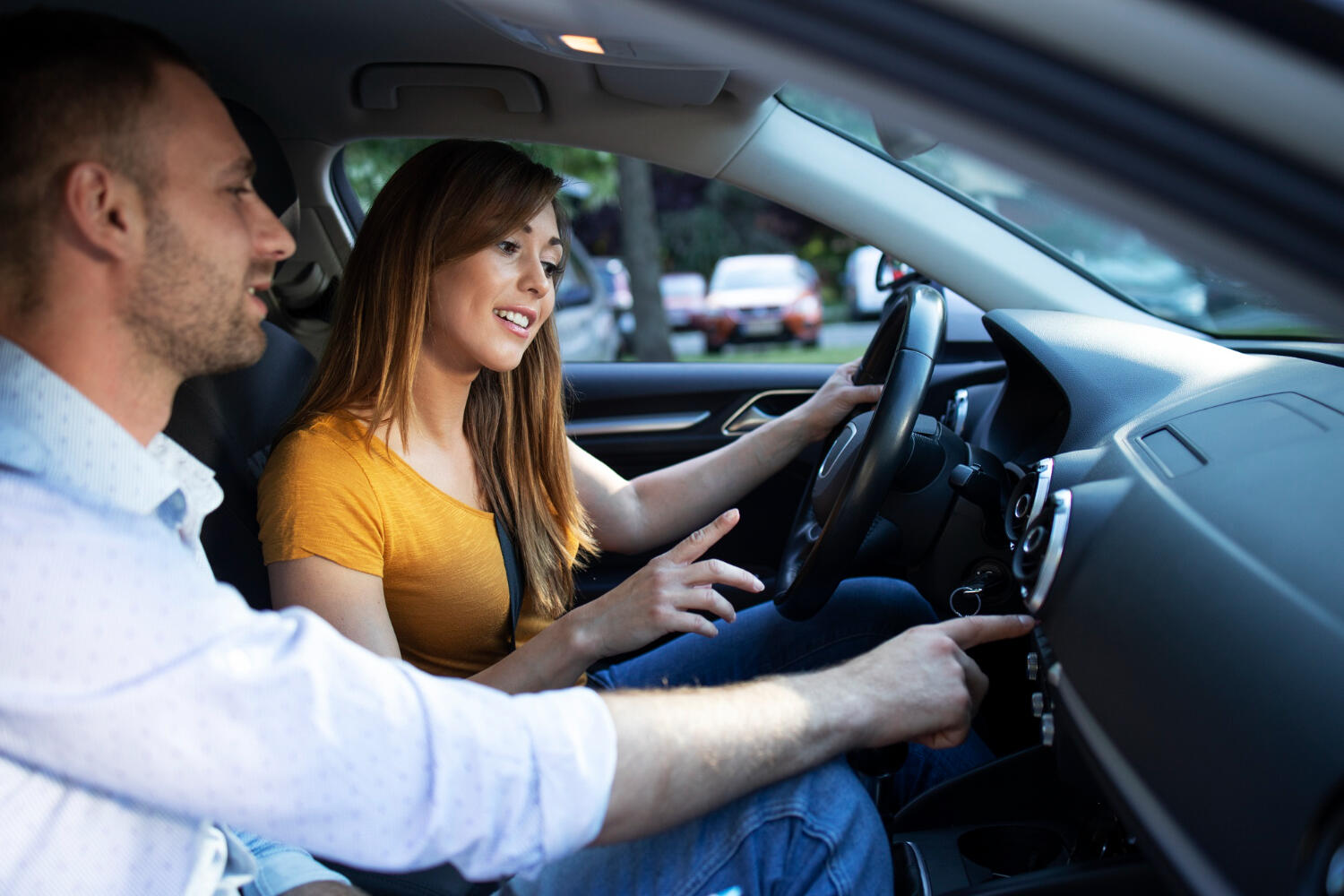 Two people inside a car, with the driver holding the steering wheel and the passenger pointing at the dashboard controls.