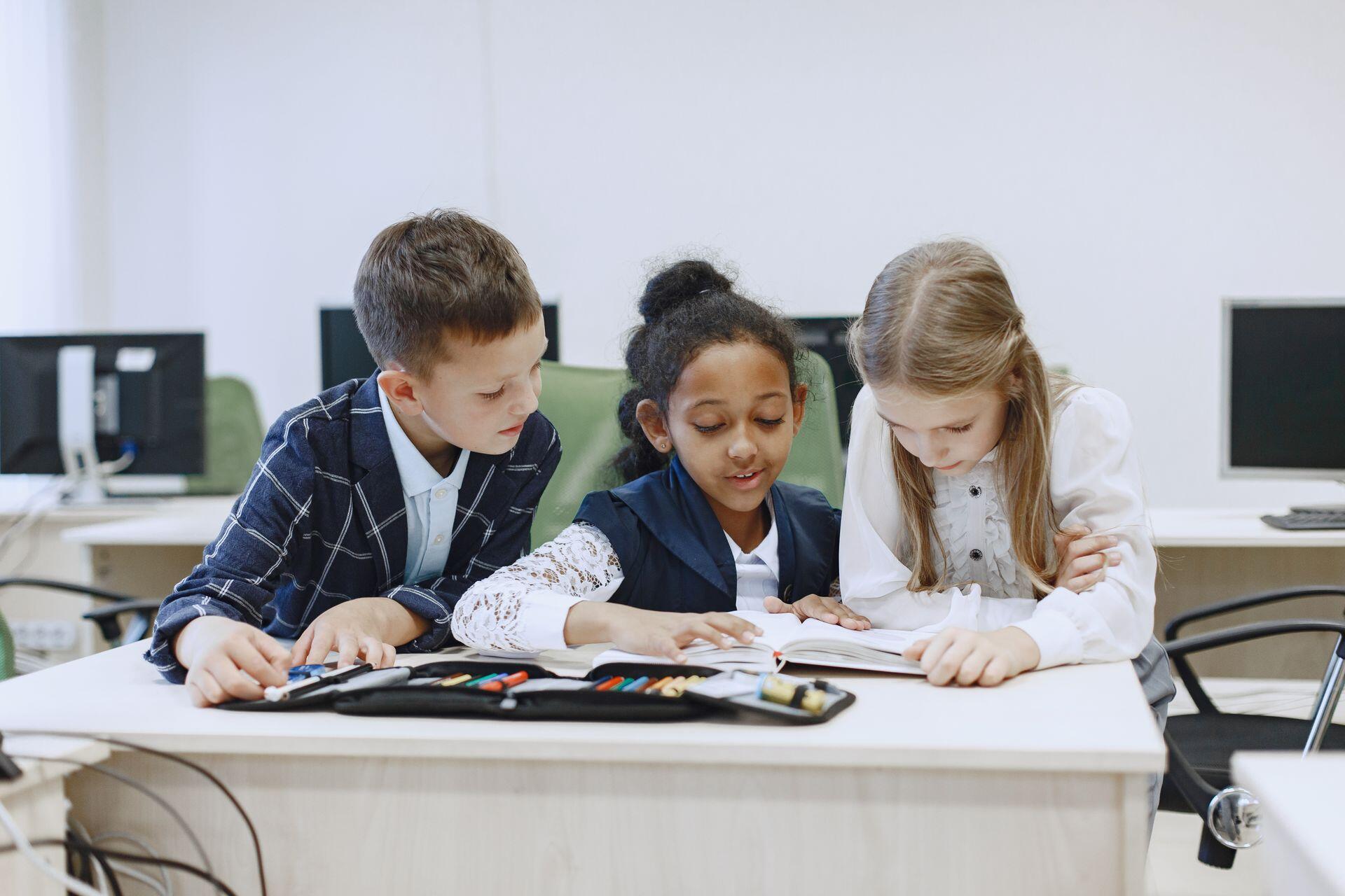 Three young students in uniforms huddle around a book at a desk with a pencil case.