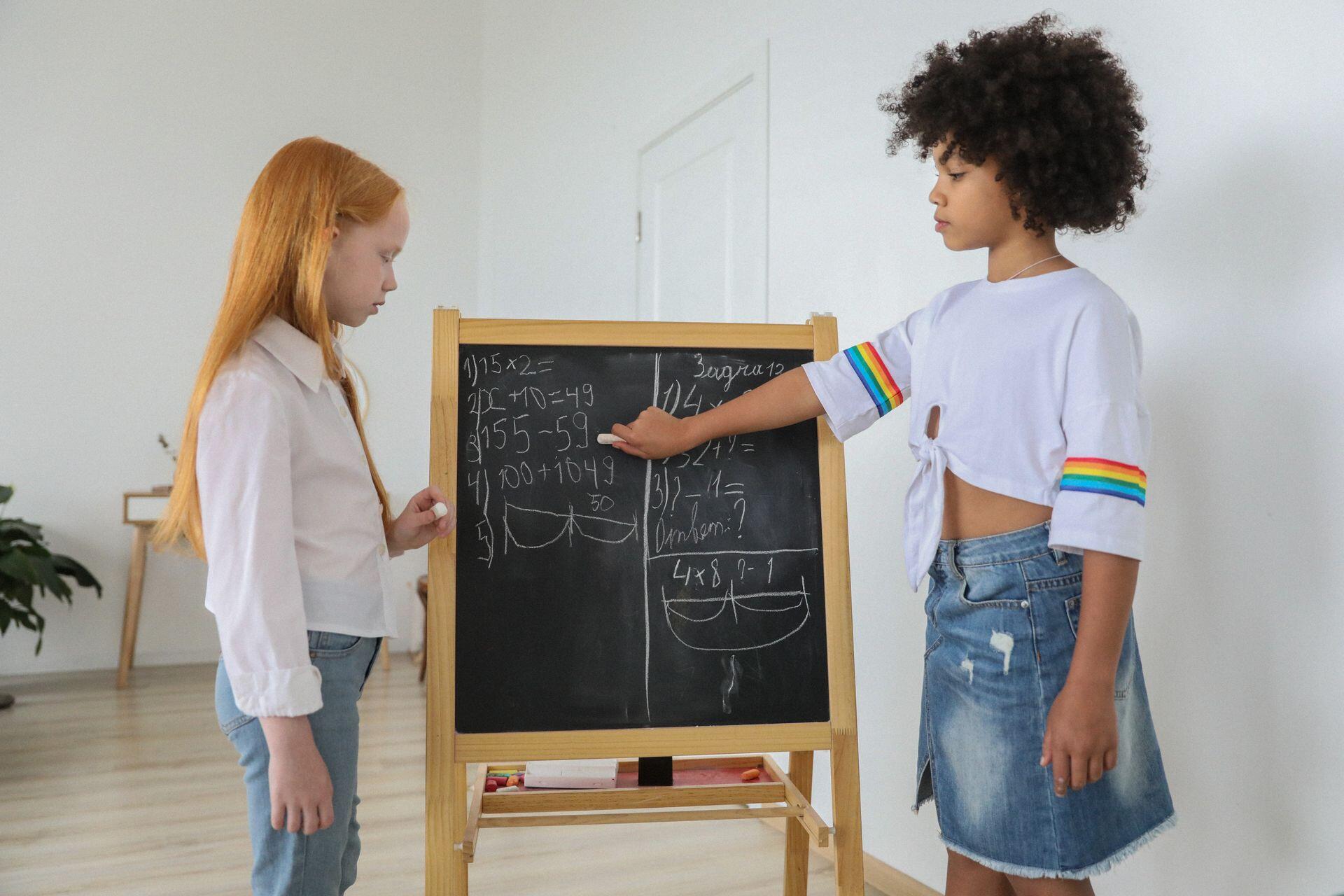 Two girls solve math problems on a chalkboard in a bright room, one pointing to the board, both dressed casually.