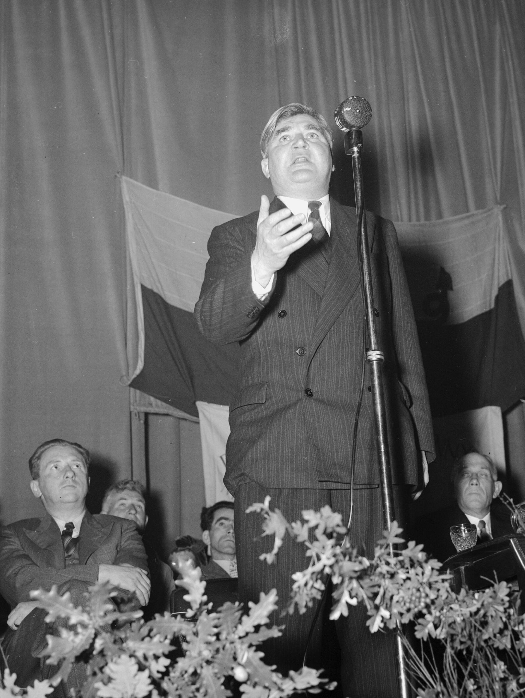 A man in a pinstripe suit and tie gestures while speaking at a microphone on a stage, with an audience seated behind him.