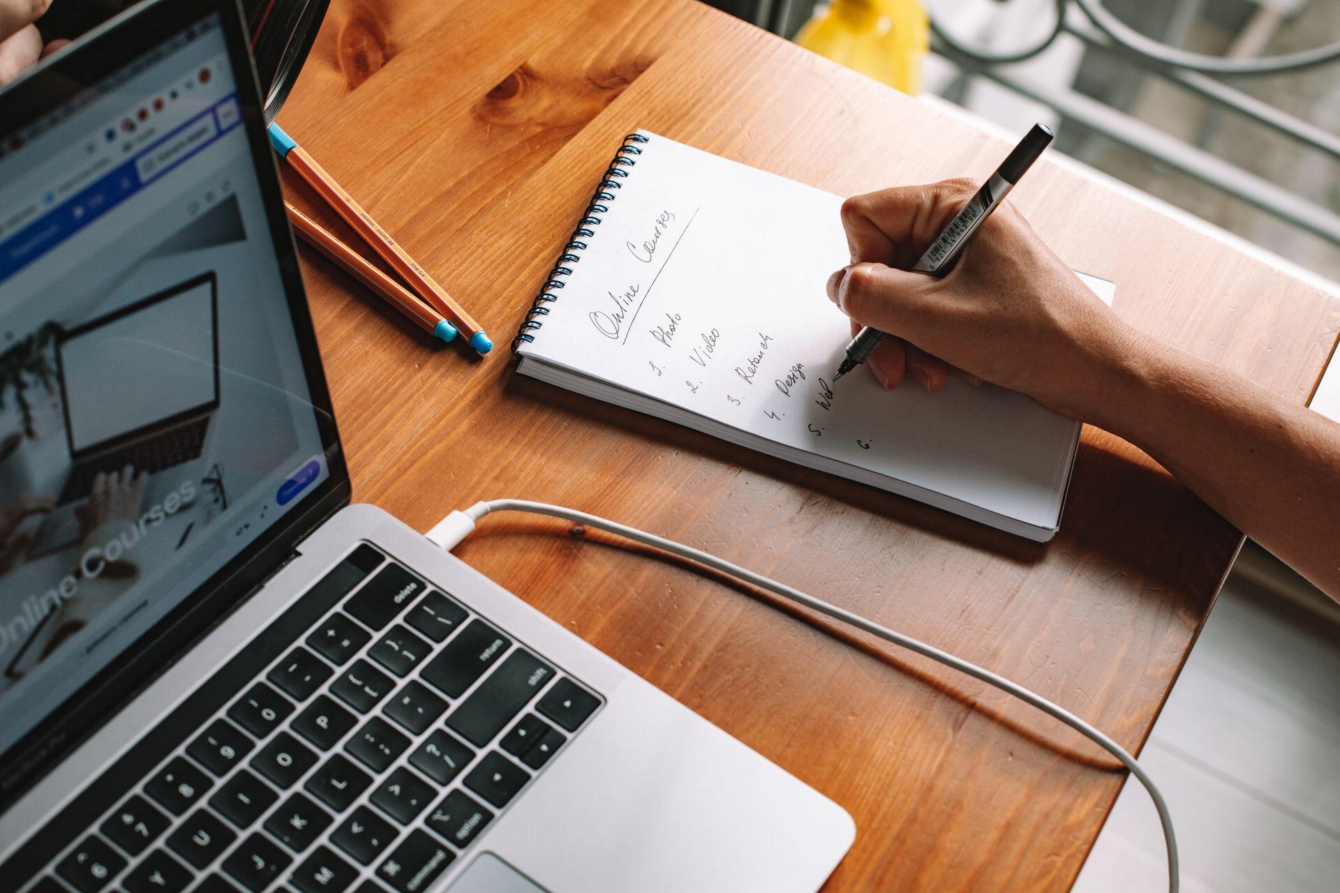 Person writing in notebook with mac computer next to them