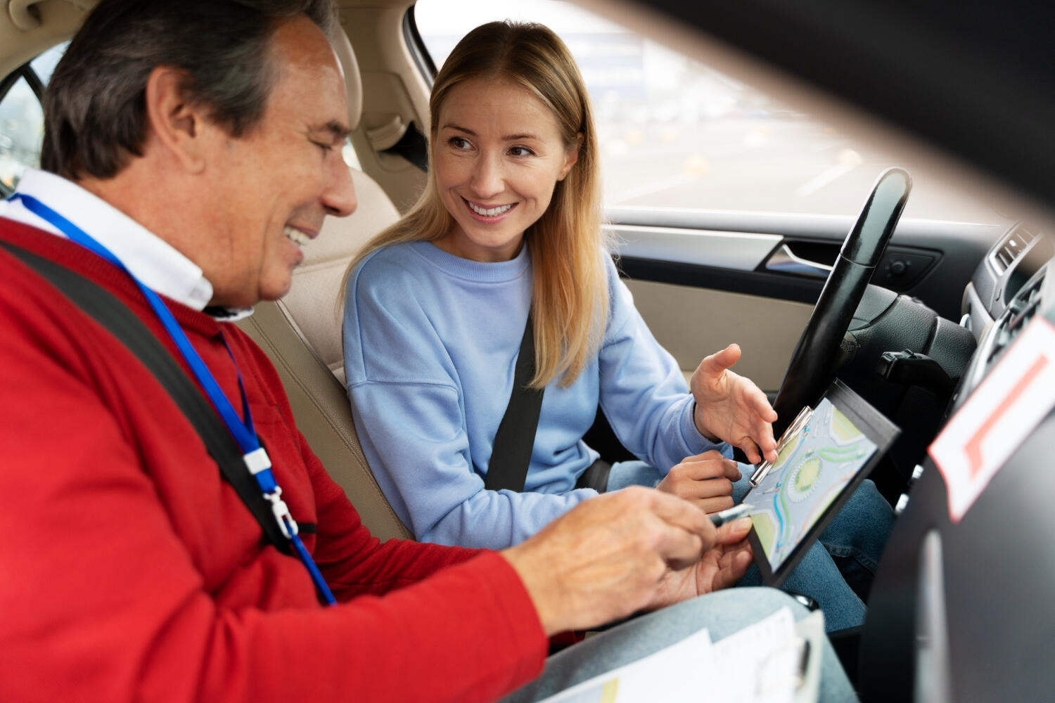 Driving instructor showing a female student a map on a clipboard inside a car during a driving lesson.