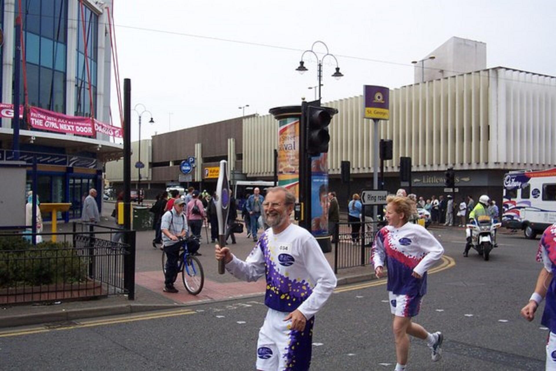 A jogger wearing a white and purple running outfit holds a baton aloft while smiling as he runs in the road, followed by another runner dressed the same way.