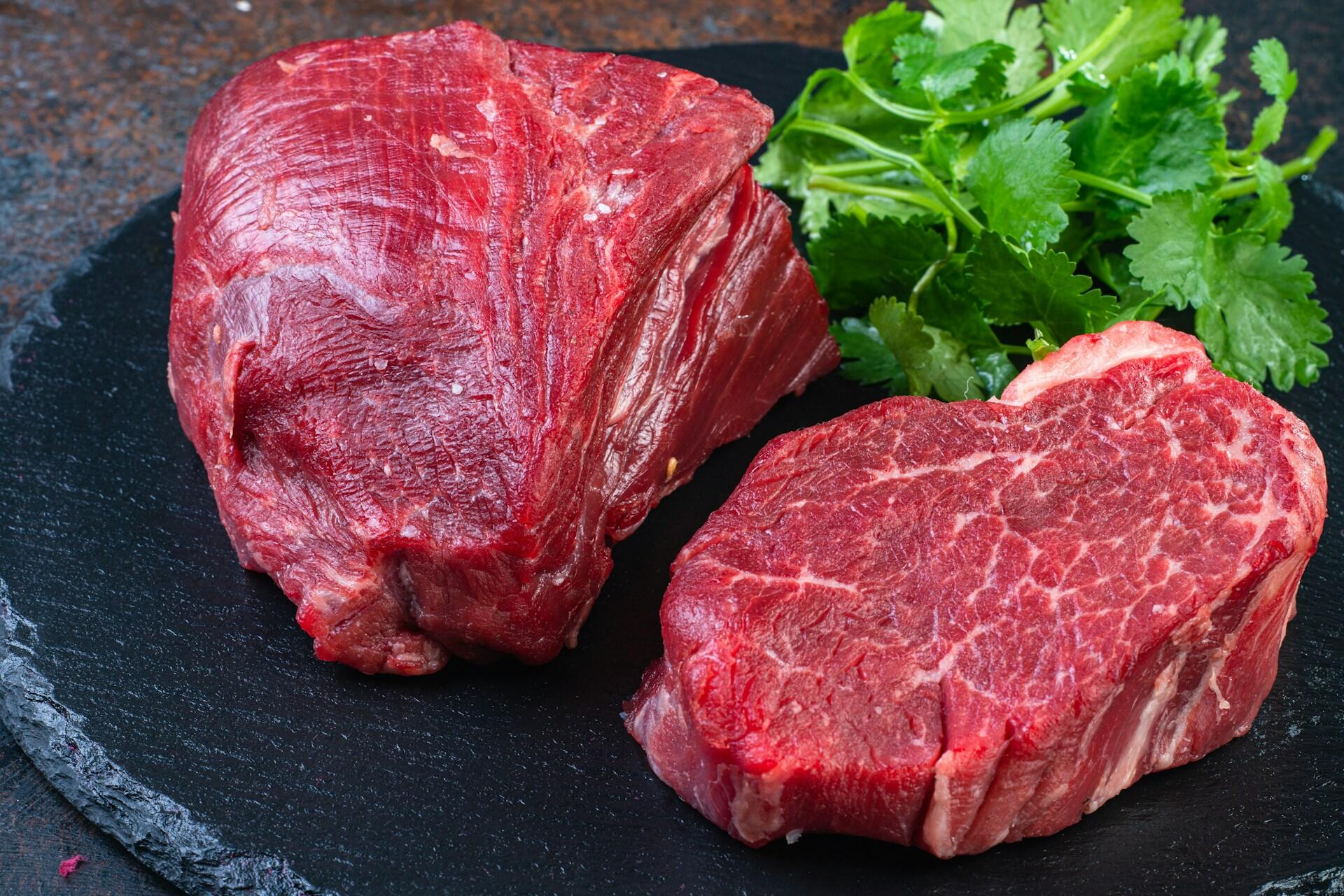 Two pieces of raw beef on a black plate with green herbs.