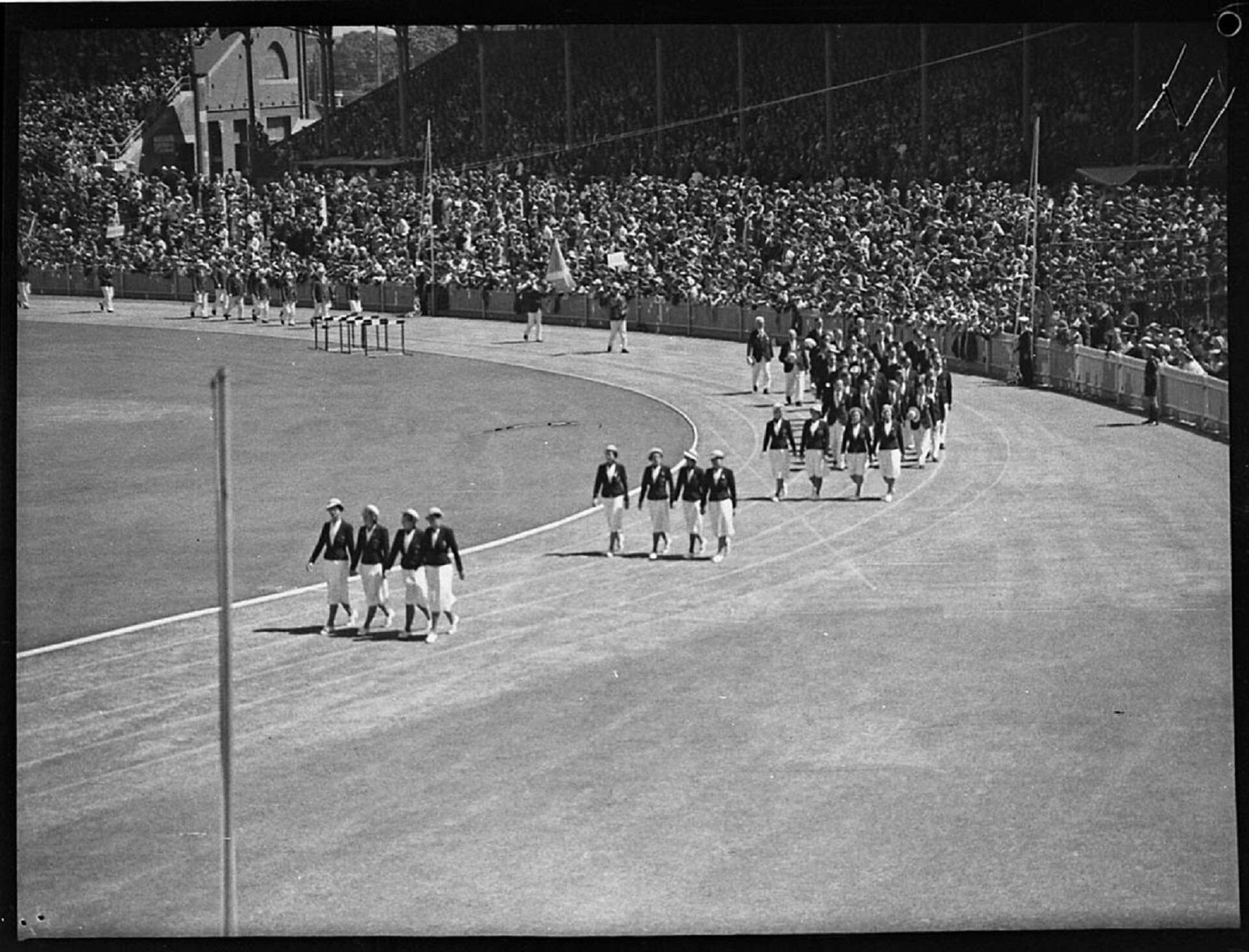 Formations of people in formal uniforms parade on a sports field, in front of full spectator stands. 