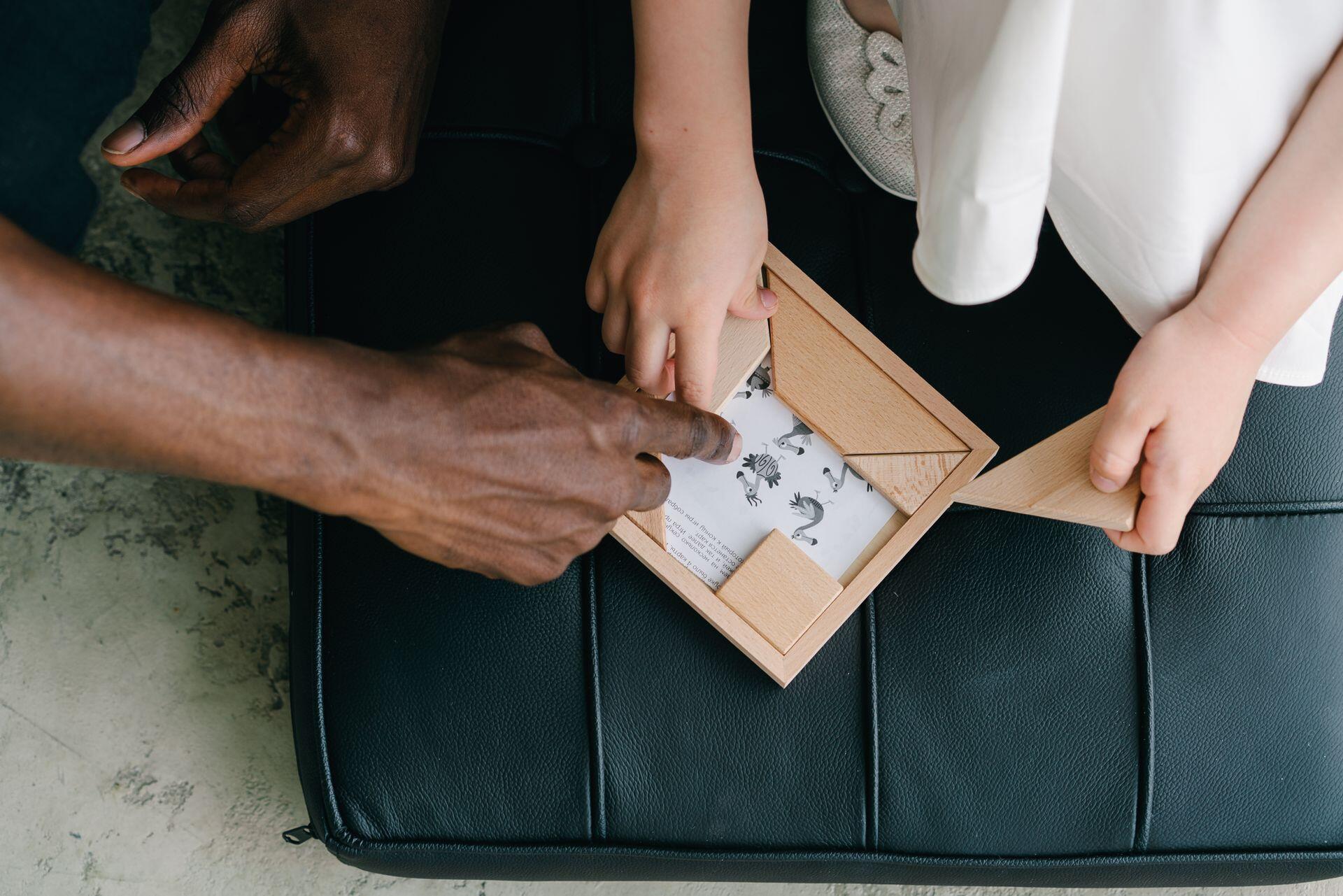 Adult and child hands assembling a wooden tangram puzzle on a black leather cushion.